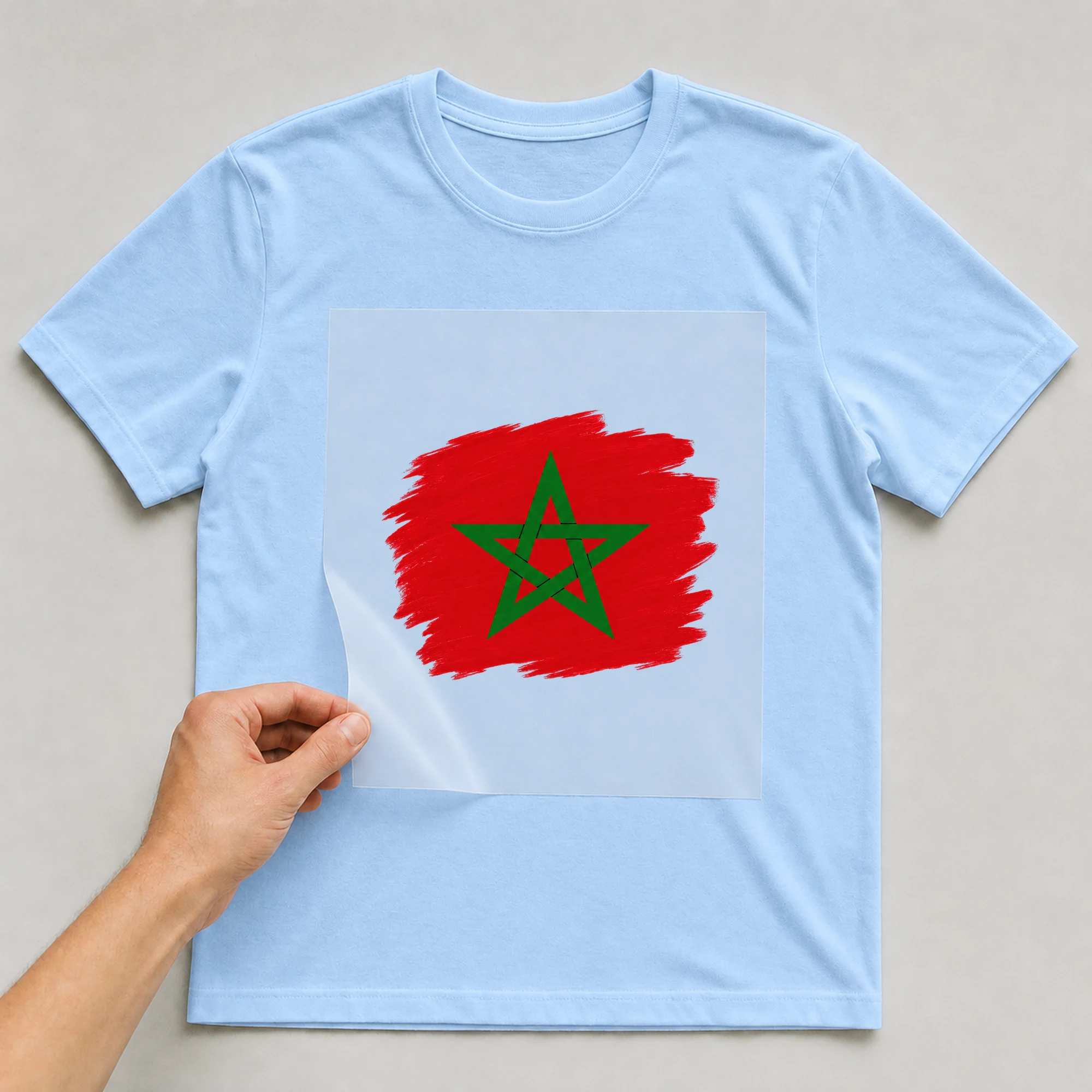 Bügelbild „Flagge Marokko im Pinselstrich“ auf babyblauem T-Shirt, Folie wird aufgelegt