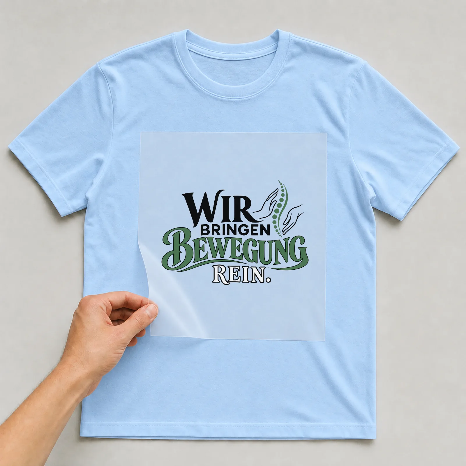 Bügelbild „Wir bringen Bewegung rein“ auf hellblauem T-Shirt, Folie wird aufgelegt