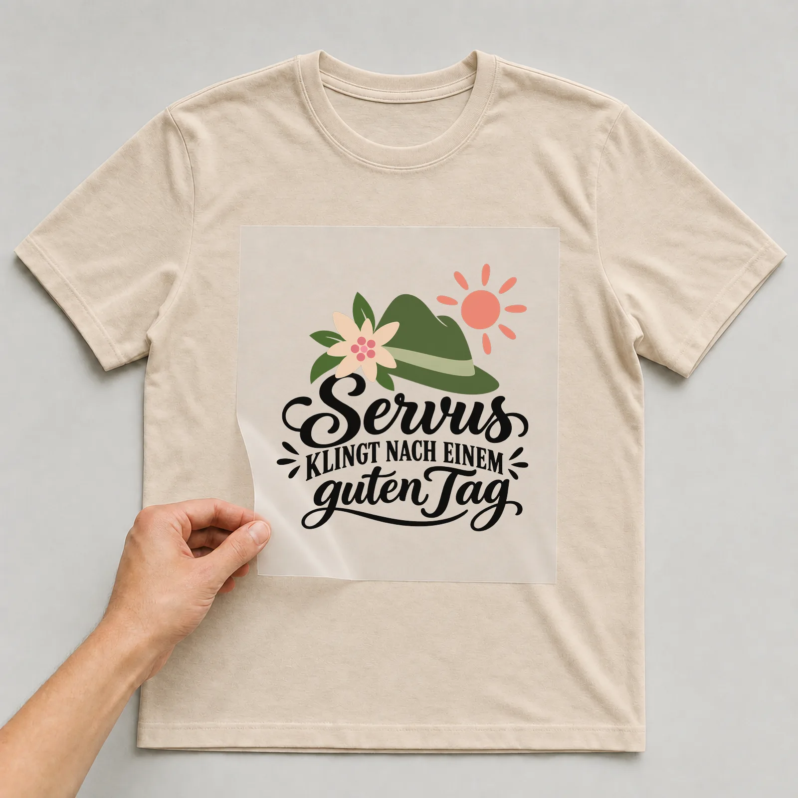 Bügelbild „Servus KLINGT NACH EINEM guten Tag“ auf beigem T-Shirt, Folie wird aufgelegt