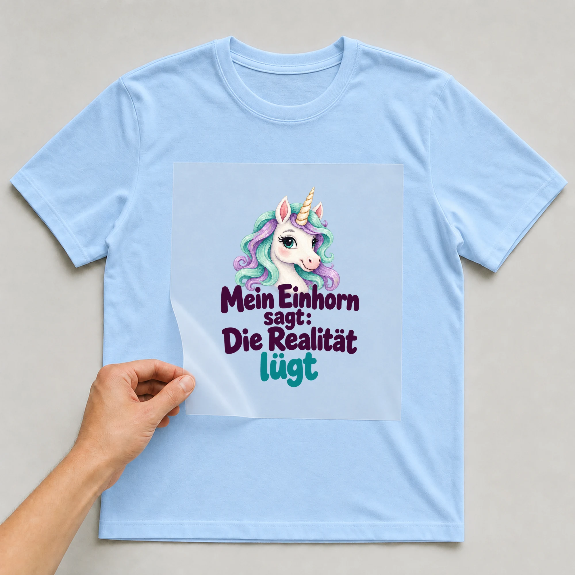 Bügelbild „Mein Einhorn sagt: Die Realität lügt“ auf babyblauem T-Shirt, Folie wird aufgelegt