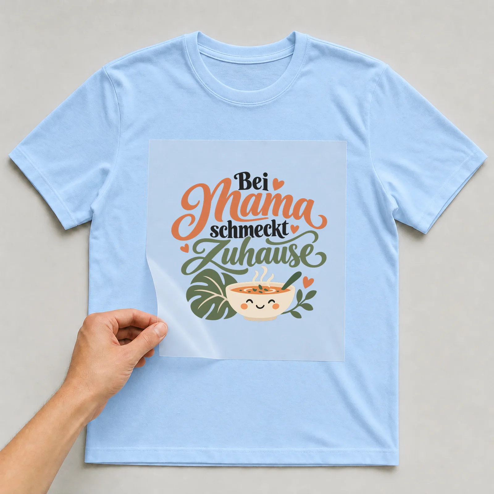 Bügelbild „Bei Mama schmeckt Zuhause“ auf hellblauem T-Shirt, Folie wird aufgelegt