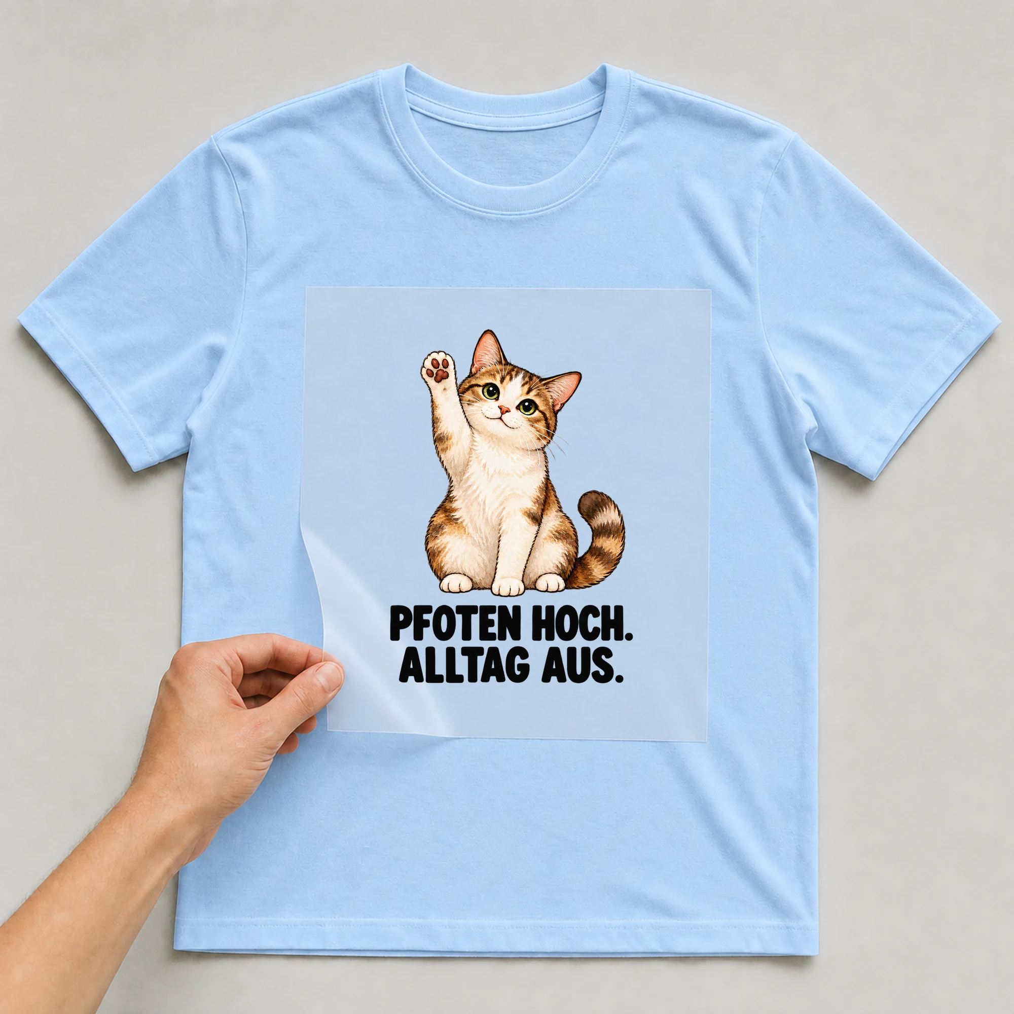 Bügelbild „PFOTEN HOCH. ALLTAG AUS.“ auf babyblauem T-Shirt, Folie wird aufgelegt