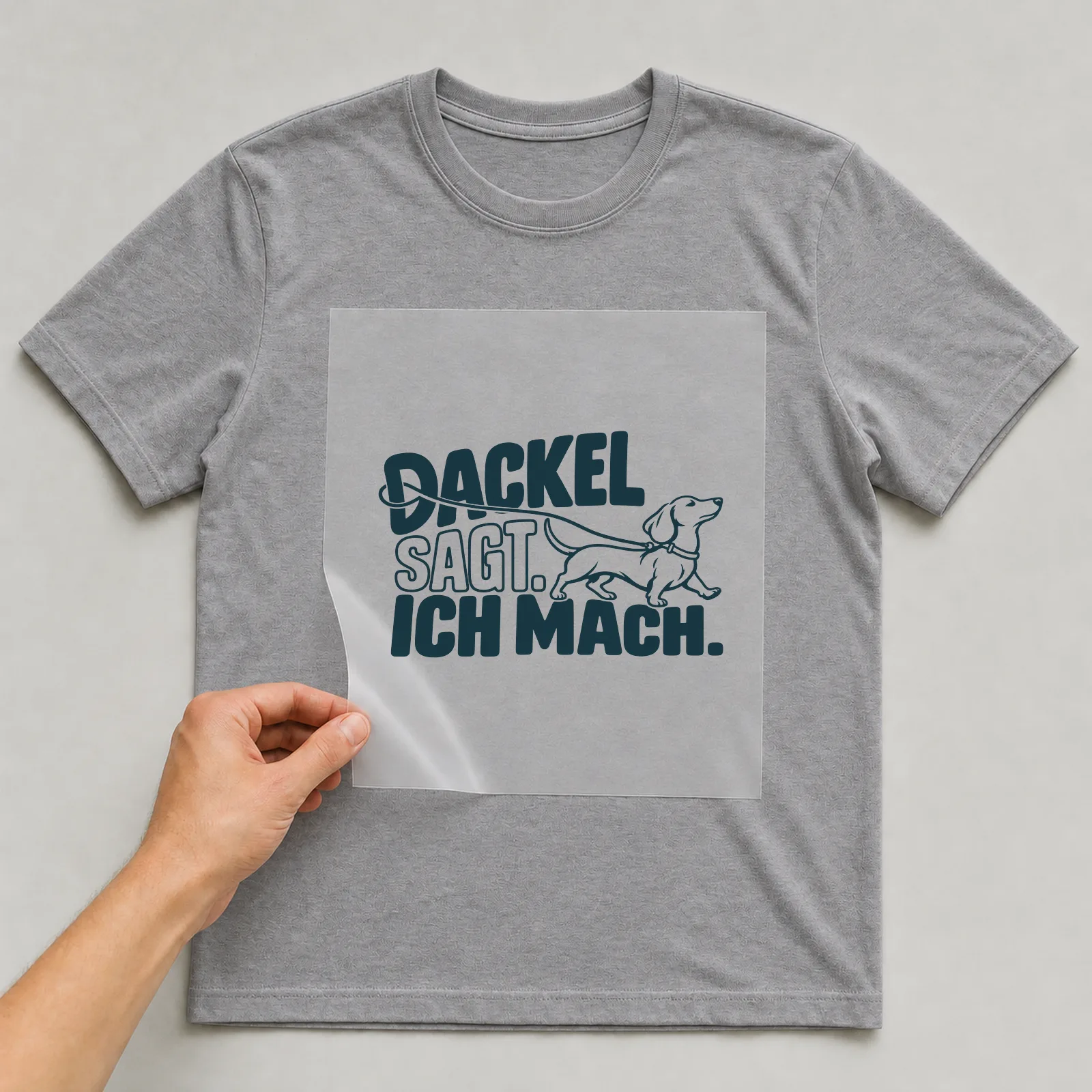 Bügelbild „Dackel sagt. Ich mach.“ auf grauem T-Shirt, Folie wird aufgelegt