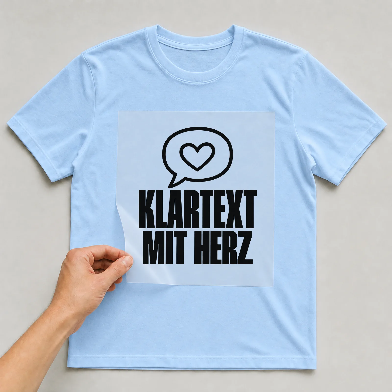 Bügelbild „Klartext mit Herz“ auf hellblauem T-Shirt, Folie wird aufgelegt