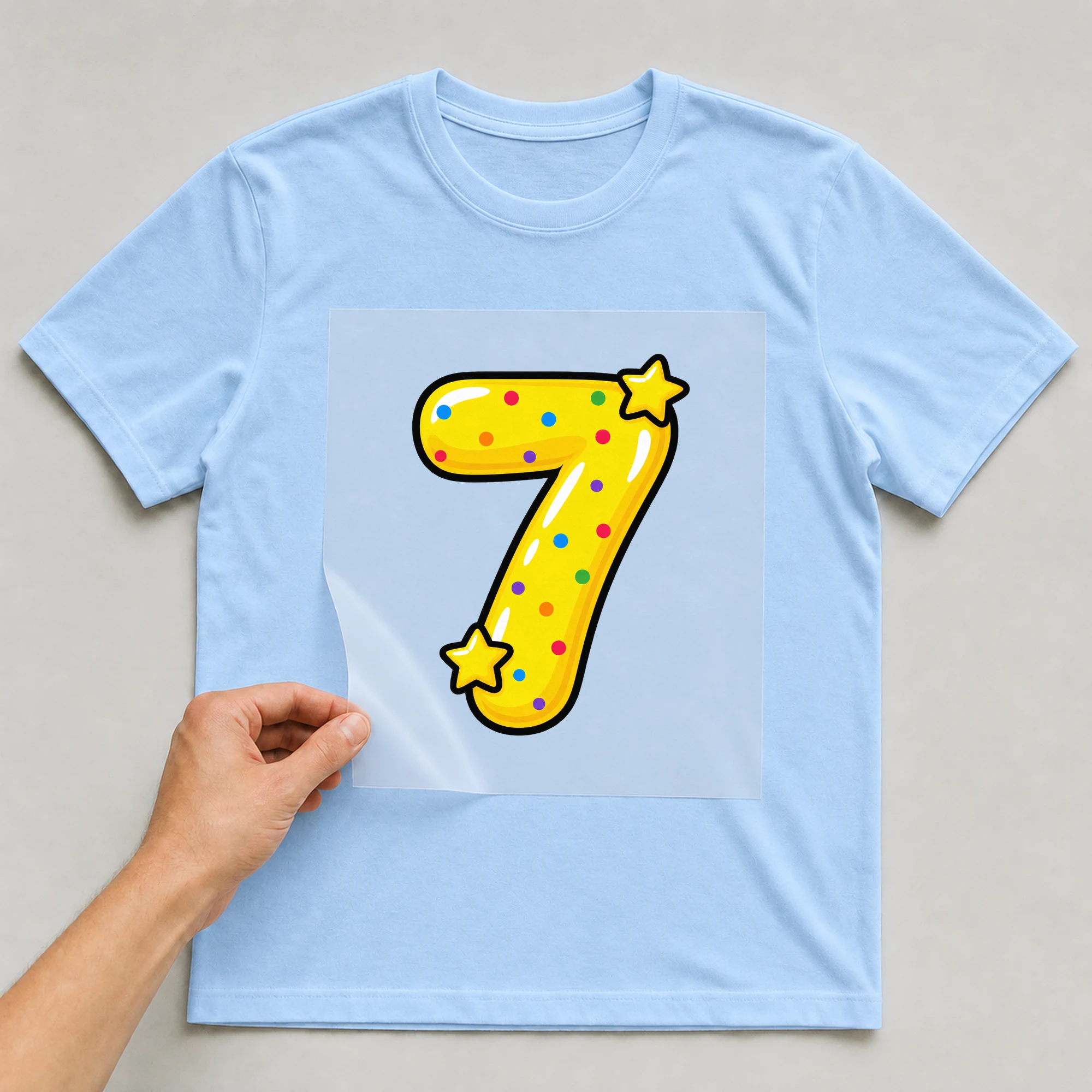 Bügelbild „Zahl 7 im Comic-Ballon-Stil“ auf babyblauem T-Shirt, Folie wird aufgelegt