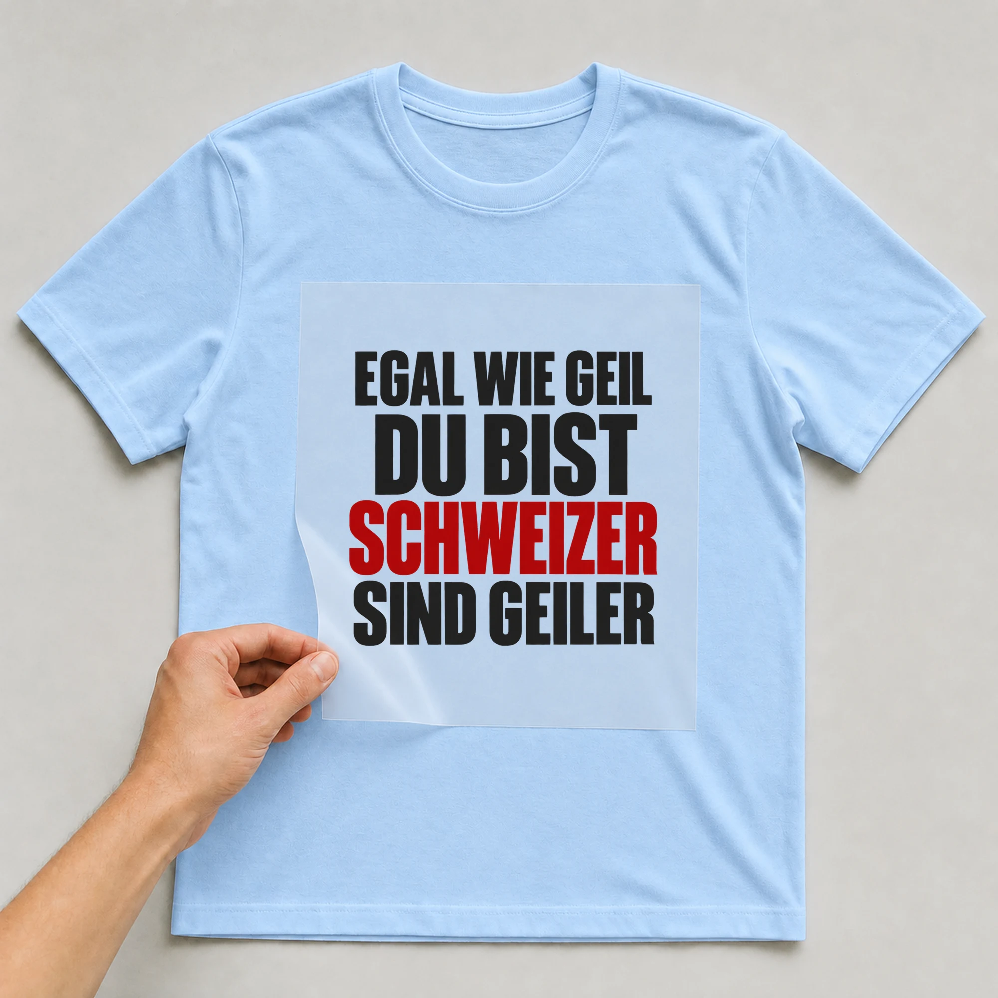 Bügelbild „Egal wie geil du bist Schweizer sind geiler“ auf babyblauem T-Shirt, Folie wird aufgelegt