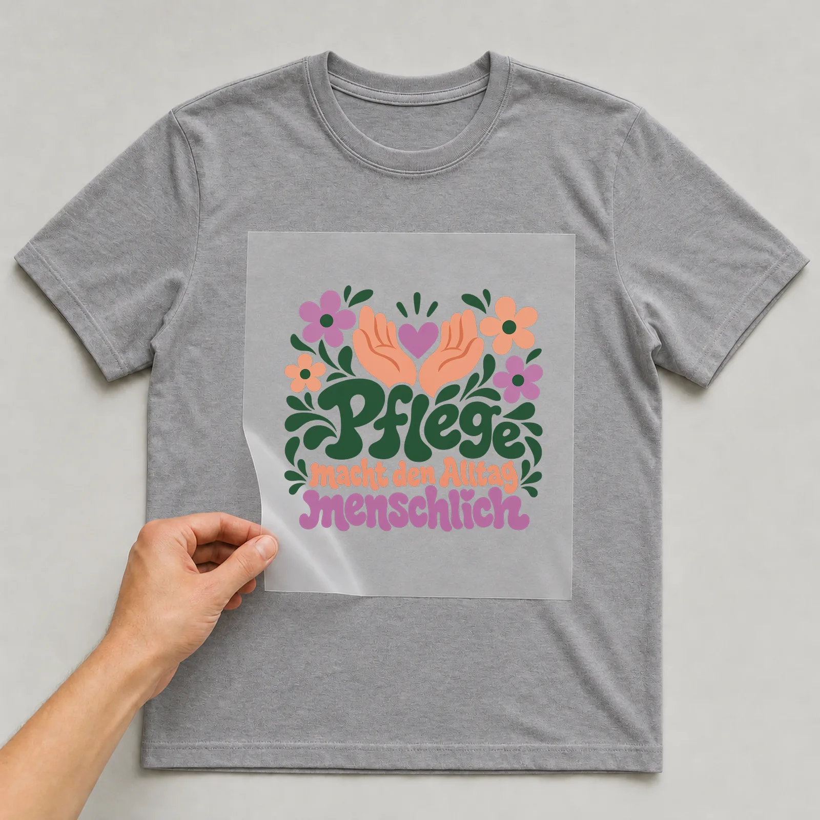 Bügelbild „Pflege macht den Alltag menschlich“ auf grauem T-Shirt, Folie wird aufgelegt