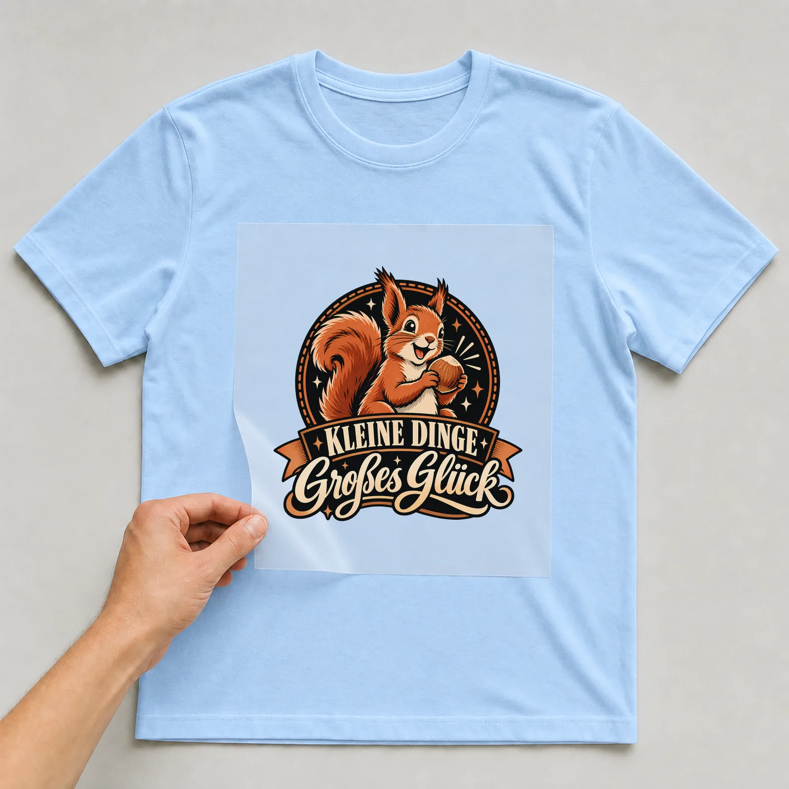 Bügelbild „Kleine Dinge Großes Glück“ (Retro-Badge) auf hellblauem T-Shirt, Folie wird aufgelegt