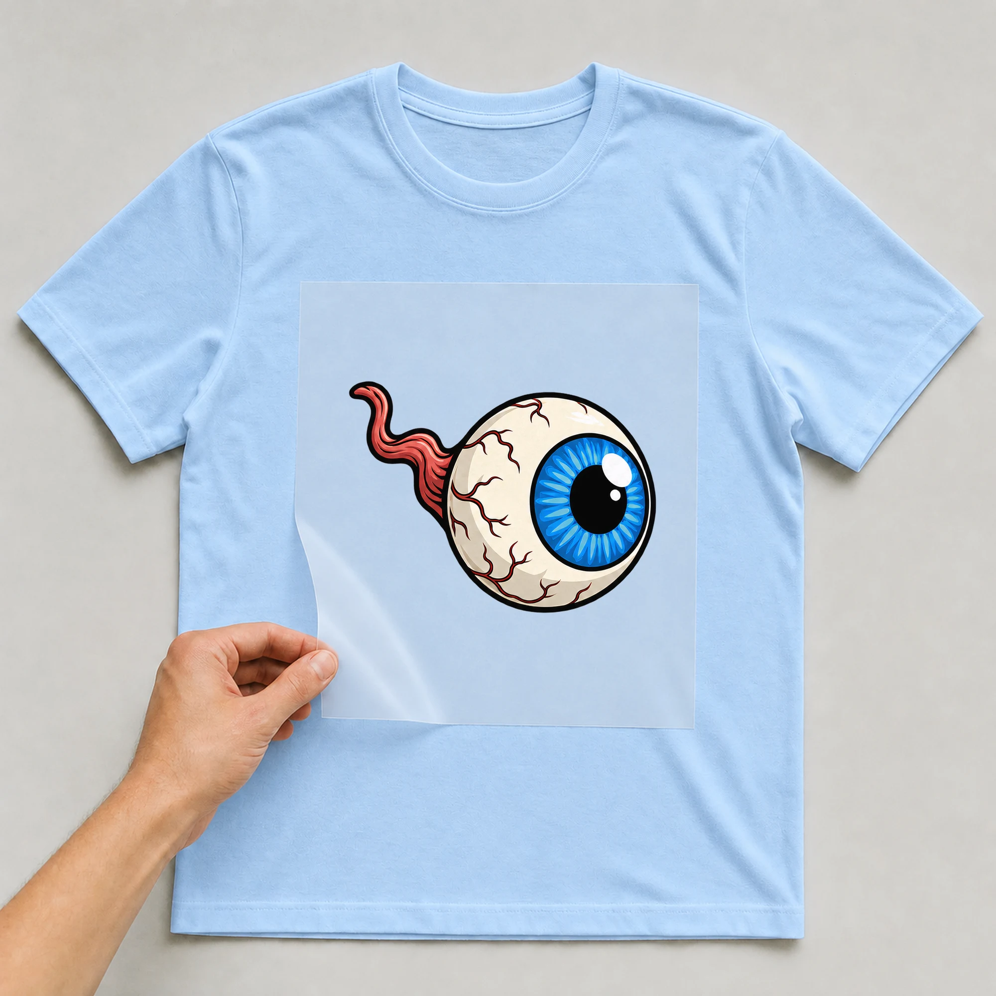 Bügelbild „Augapfel mit blauer Iris“ auf babyblauem T-Shirt, Folie wird aufgelegt
