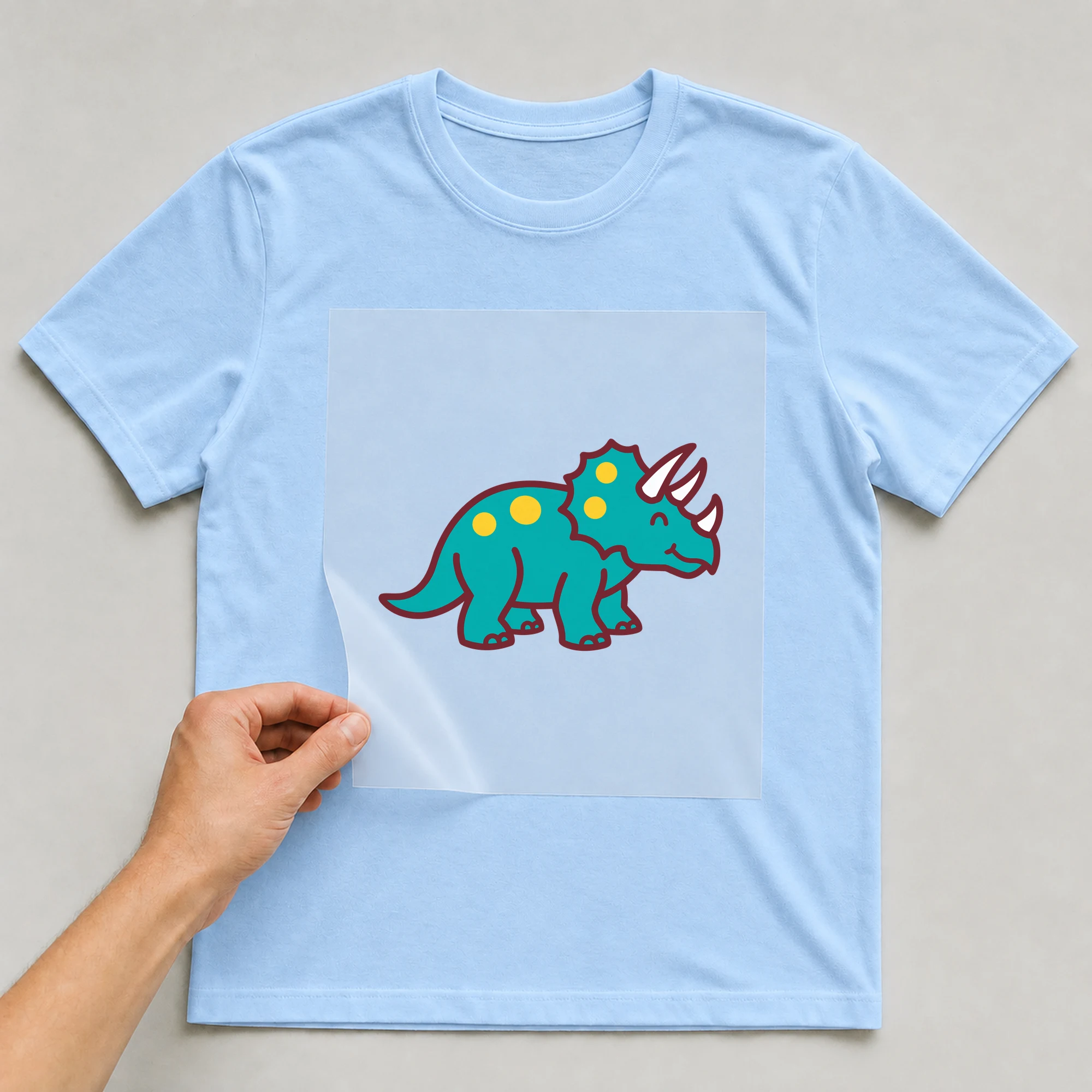 Bügelbild „Dino Triceratops“ auf babyblauem T-Shirt, Folie wird aufgelegt