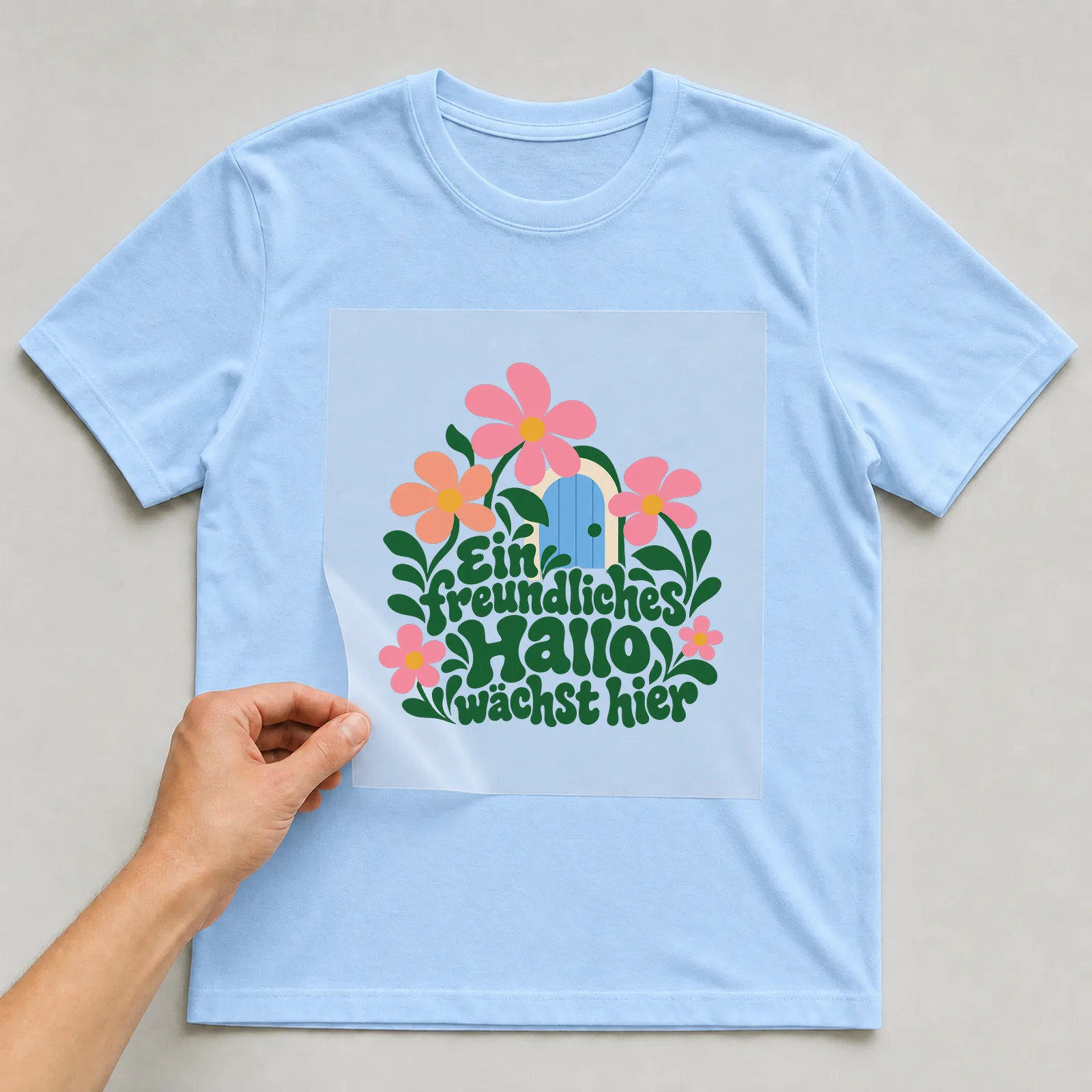 Bügelbild „Ein freundliches Hallo wächst hier“ auf hellblauem T-Shirt, Folie wird aufgelegt