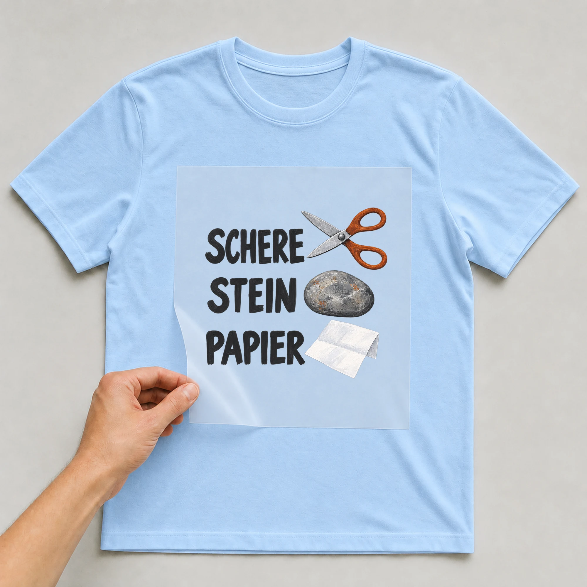 Bügelbild „Schere Stein Papier“ auf babyblauem T-Shirt, Folie wird aufgelegt