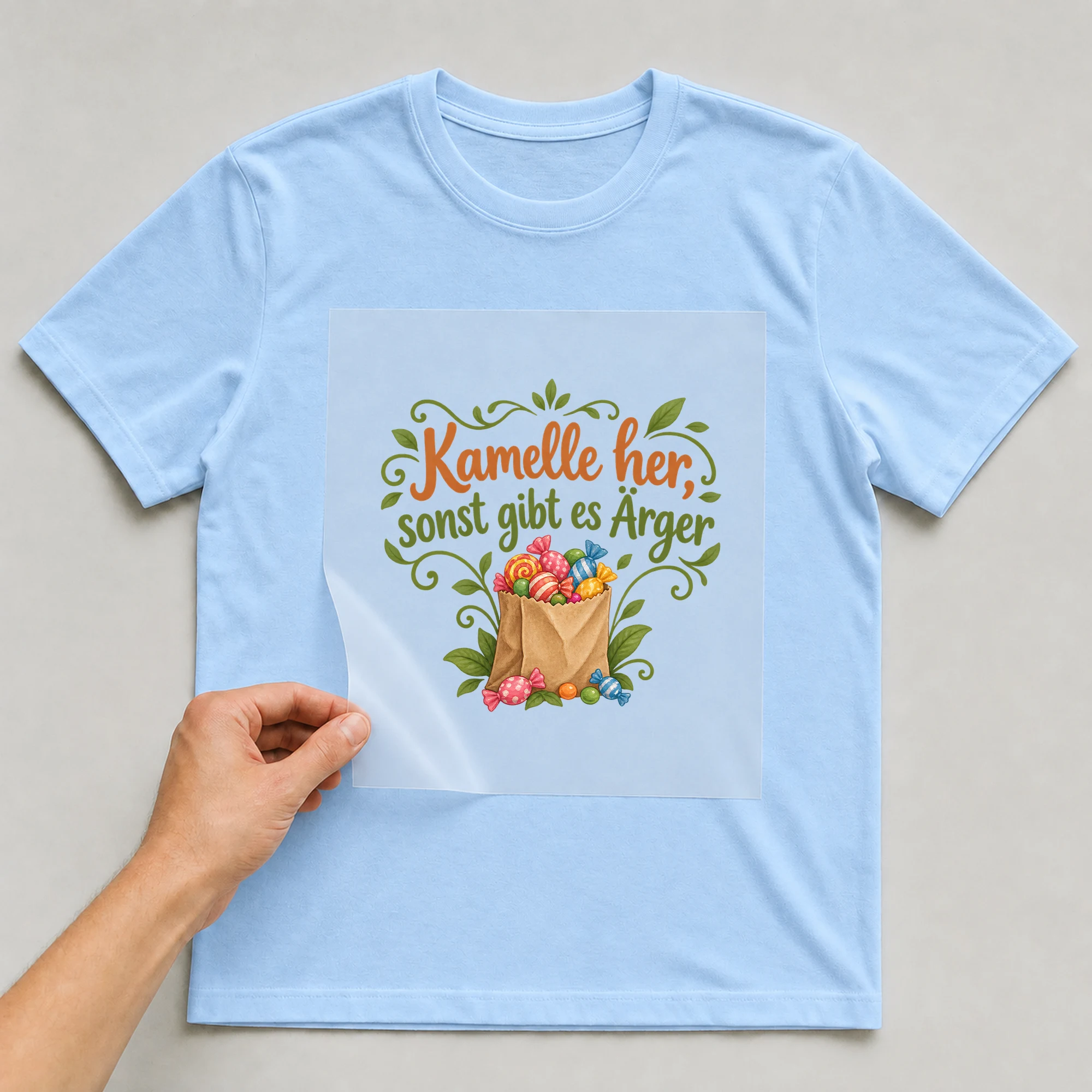 Bügelbild „Kamelle her, sonst gibt es Ärger“ auf babyblauem T-Shirt, Folie wird aufgelegt