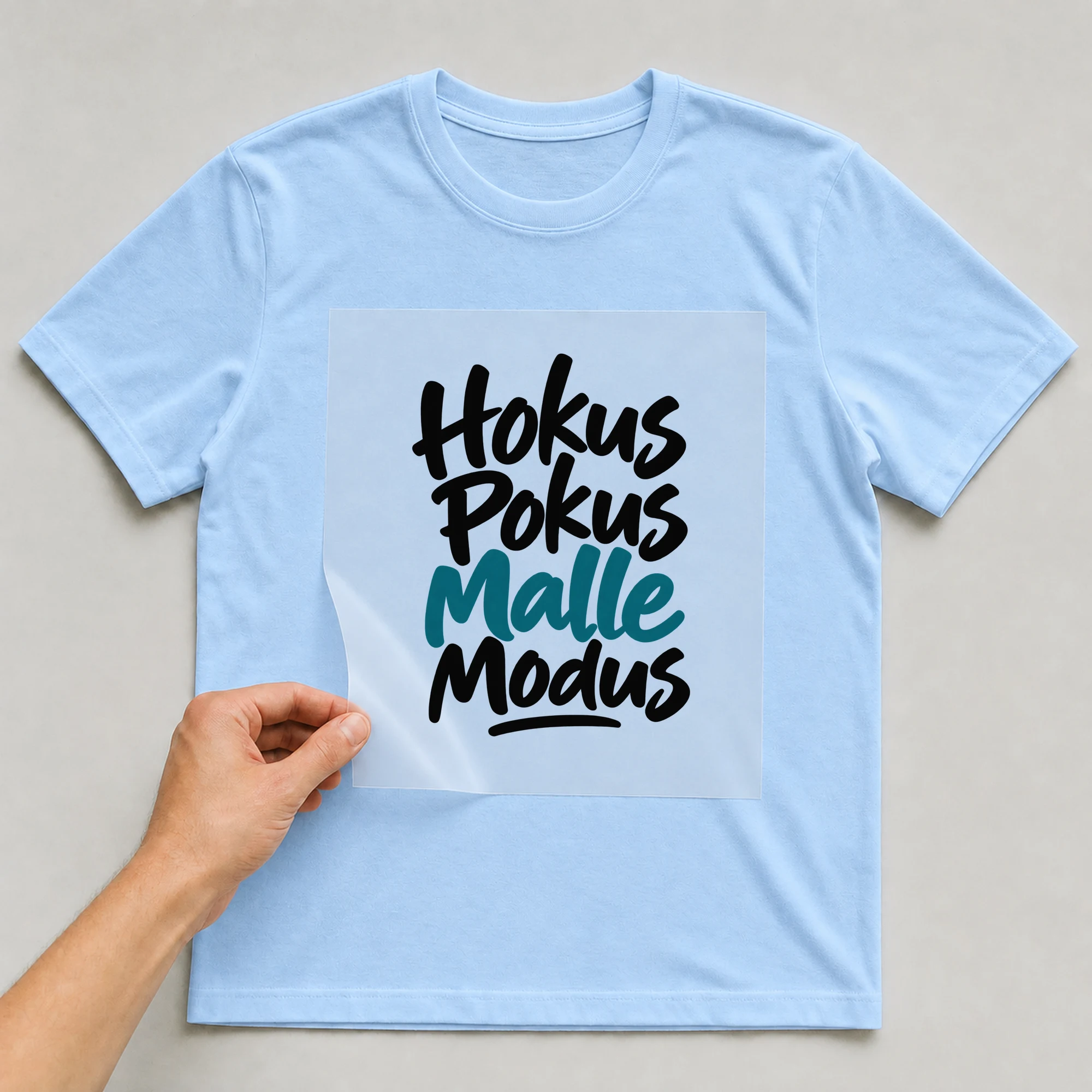 Bügelbild „Hokus Pokus Malle Modus“ auf babyblauem T-Shirt, Folie wird aufgelegt