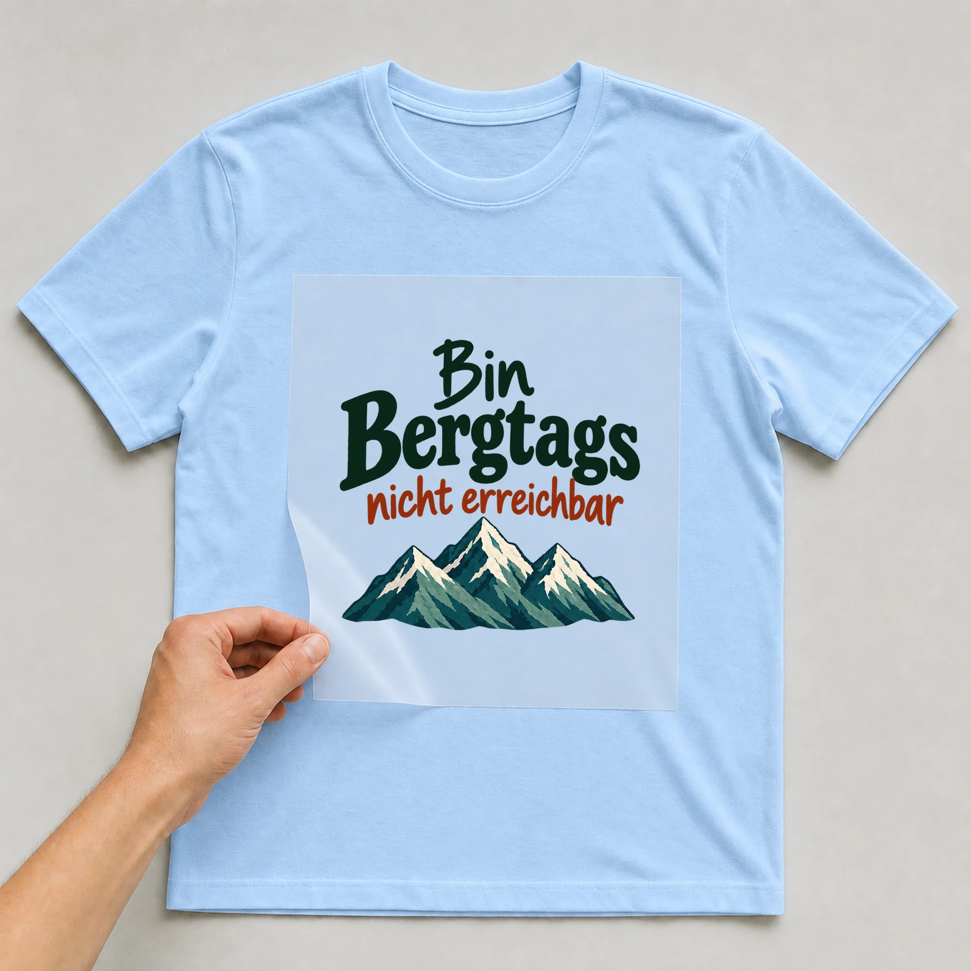 Bügelbild „Bin Bergtags nicht erreichbar“ auf babyblauem T-Shirt, Folie wird aufgelegt