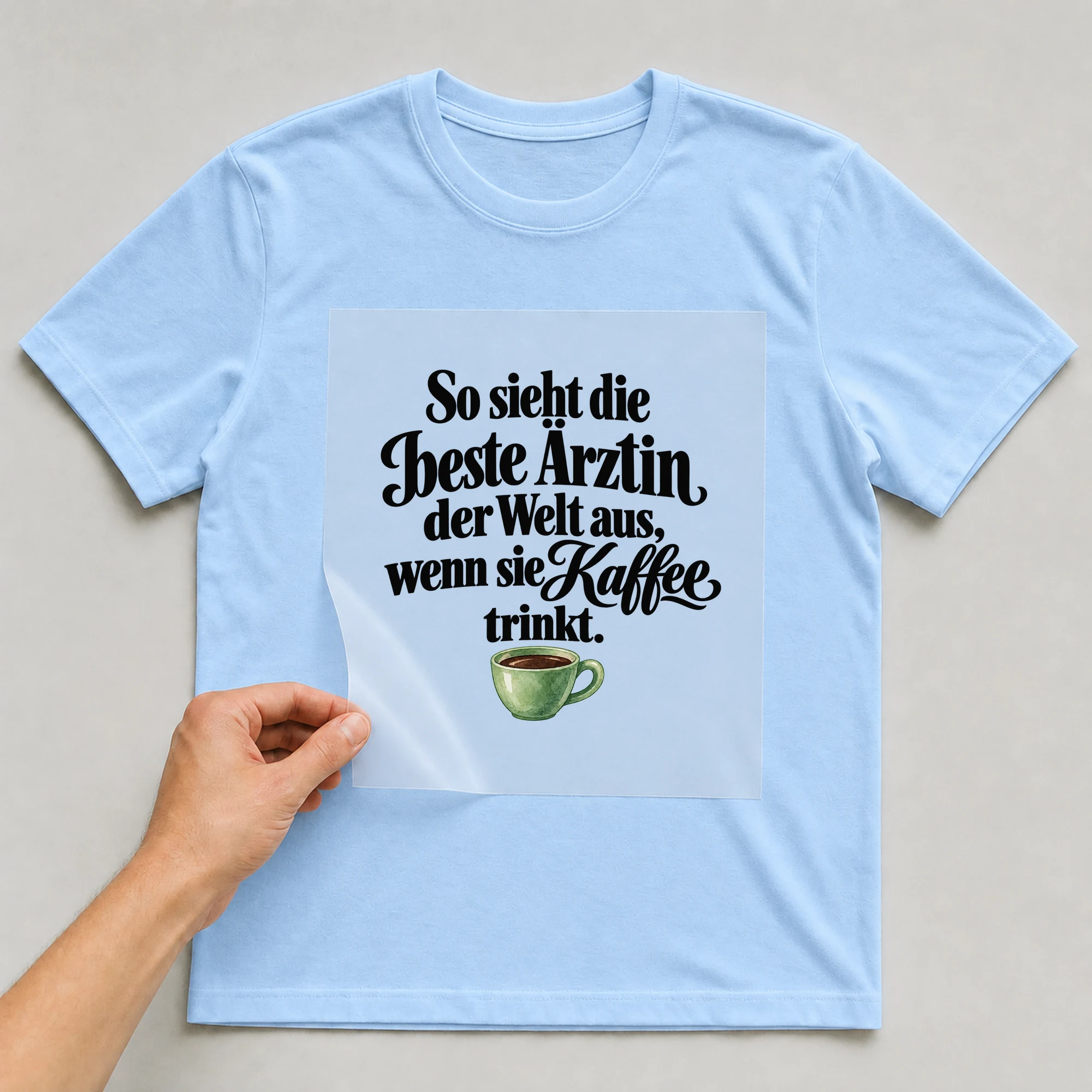 Bügelbild „So sieht die beste Ärztin der Welt aus, wenn sie Kaffee trinkt.“ auf babyblauem T-Shirt, Folie wird aufgelegt