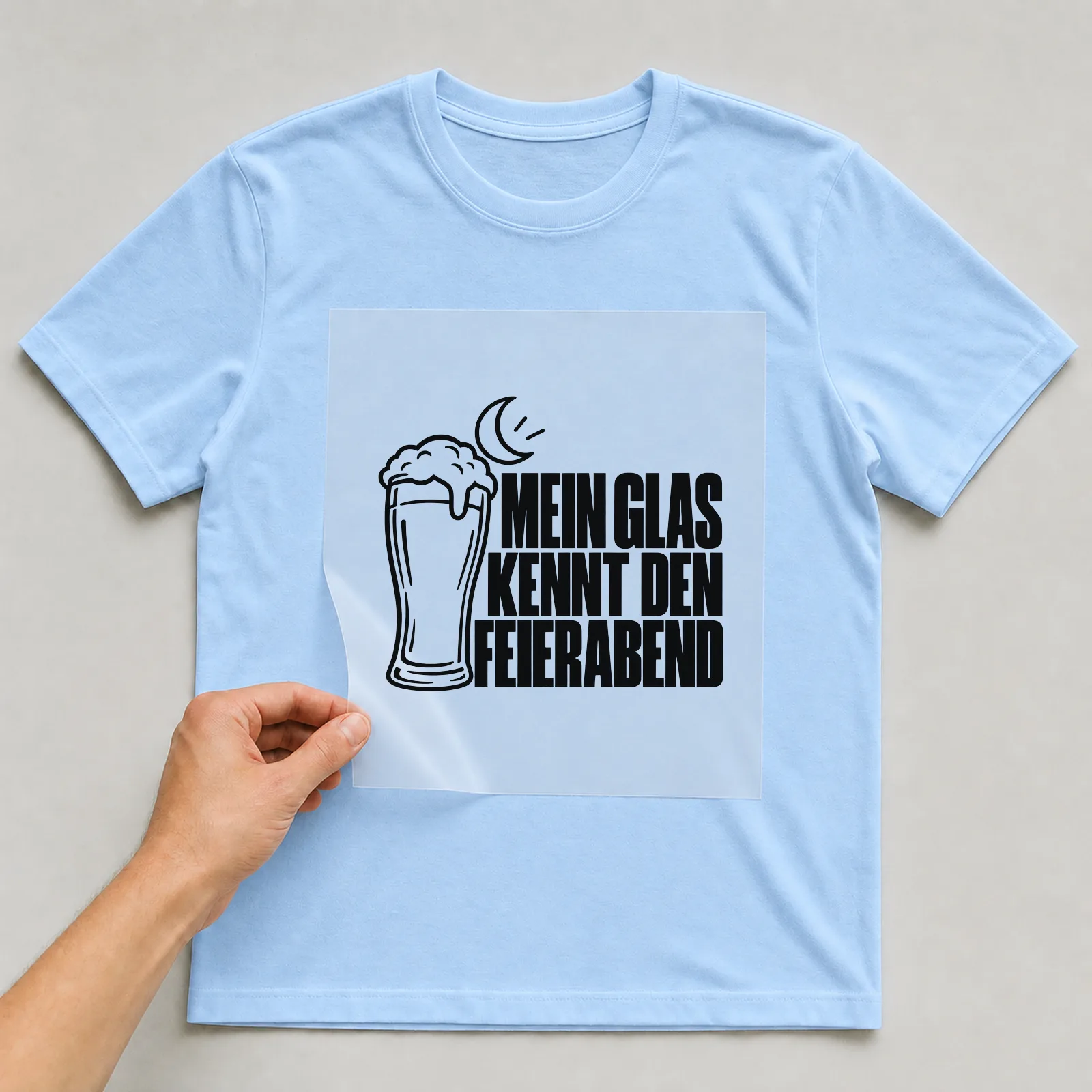 Bügelbild „Mein Glas kennt den Feierabend“ auf hellblauem T-Shirt, Folie wird aufgelegt