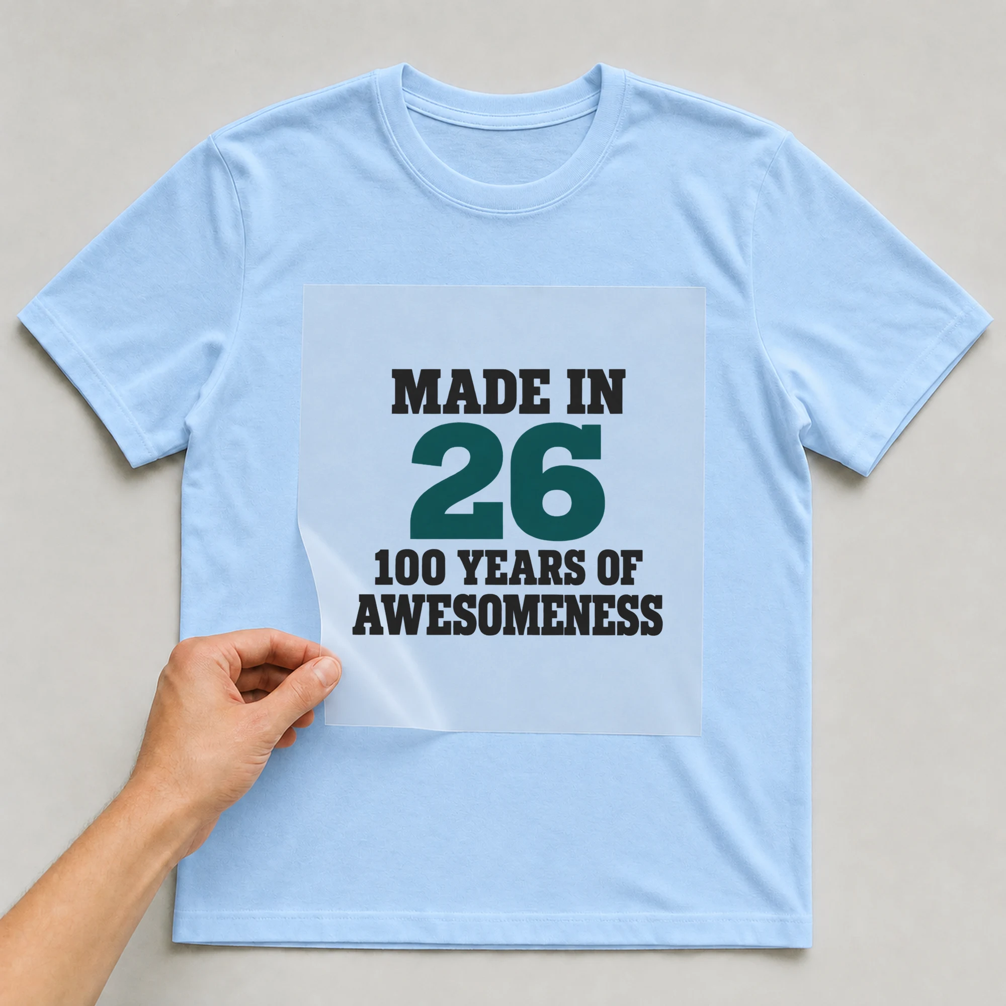 Bügelbild „Made In 26 100 Years Of Awesomeness“ auf babyblauem T-Shirt, Folie wird aufgelegt