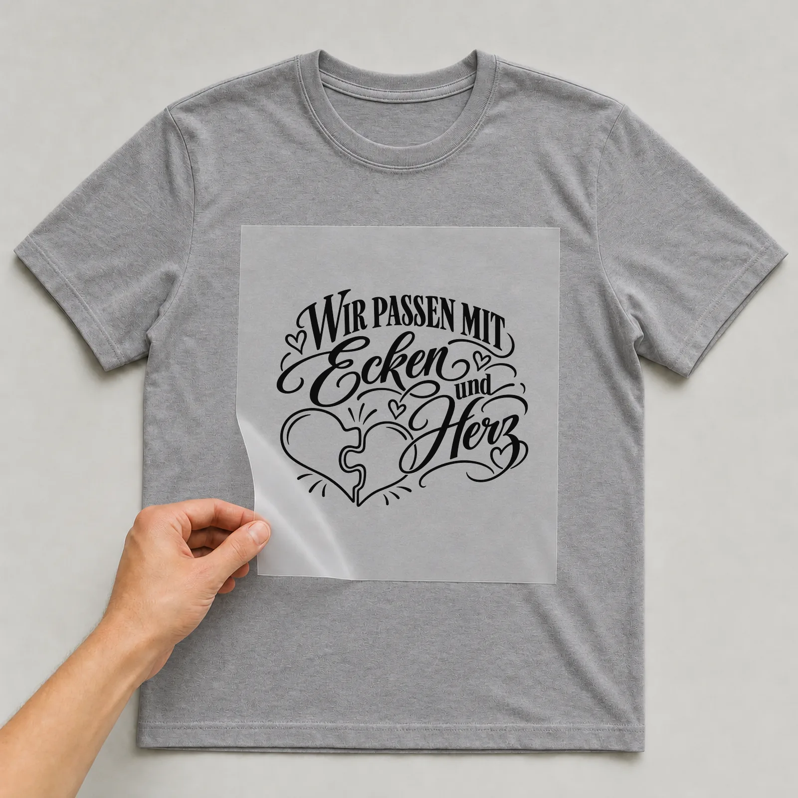 Bügelbild „Wir passen mit Ecken und Herz“ auf grauem T-Shirt, Folie wird aufgelegt