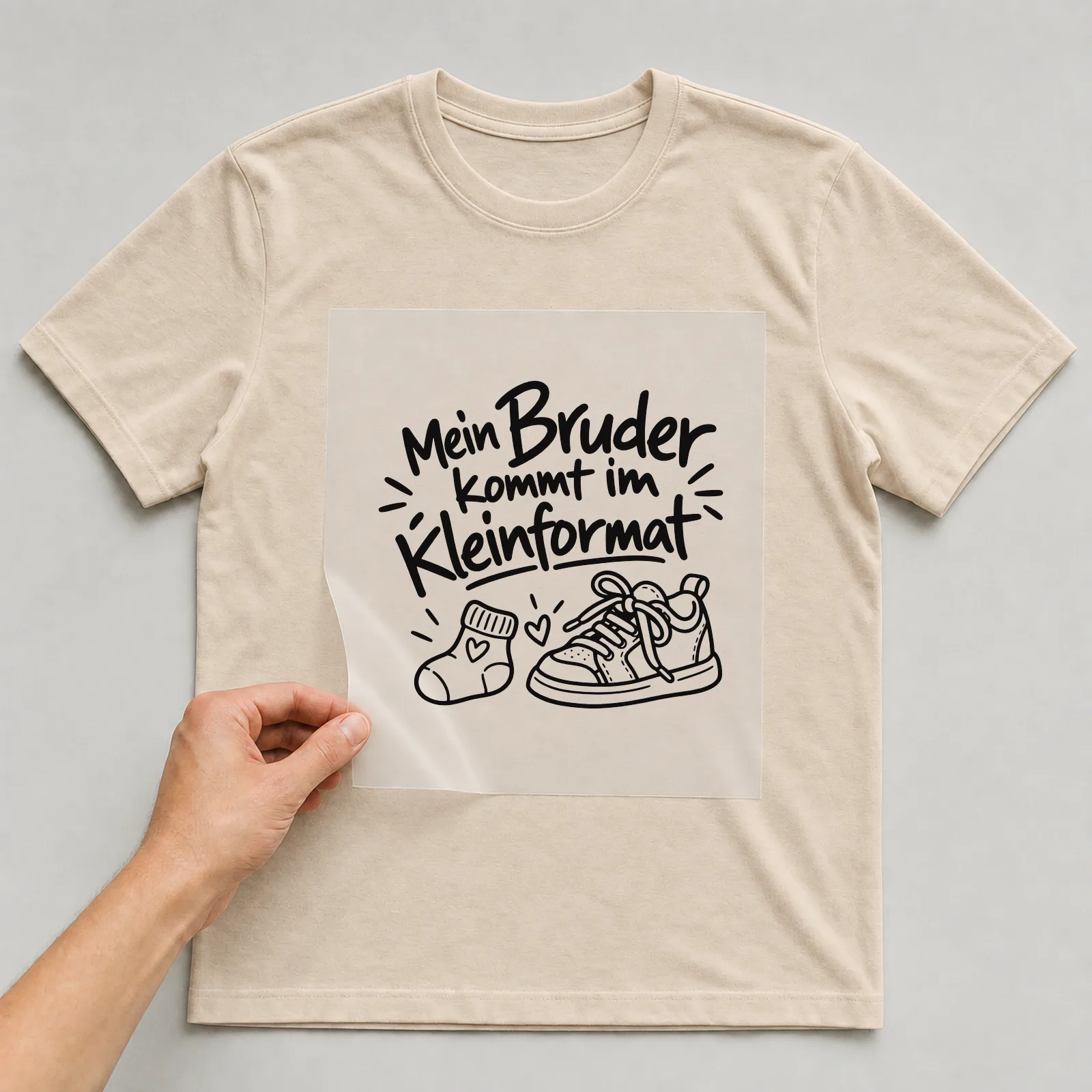 Bügelbild „Mein Bruder kommt im Kleinformat“ auf beigem T-Shirt, Folie wird aufgelegt