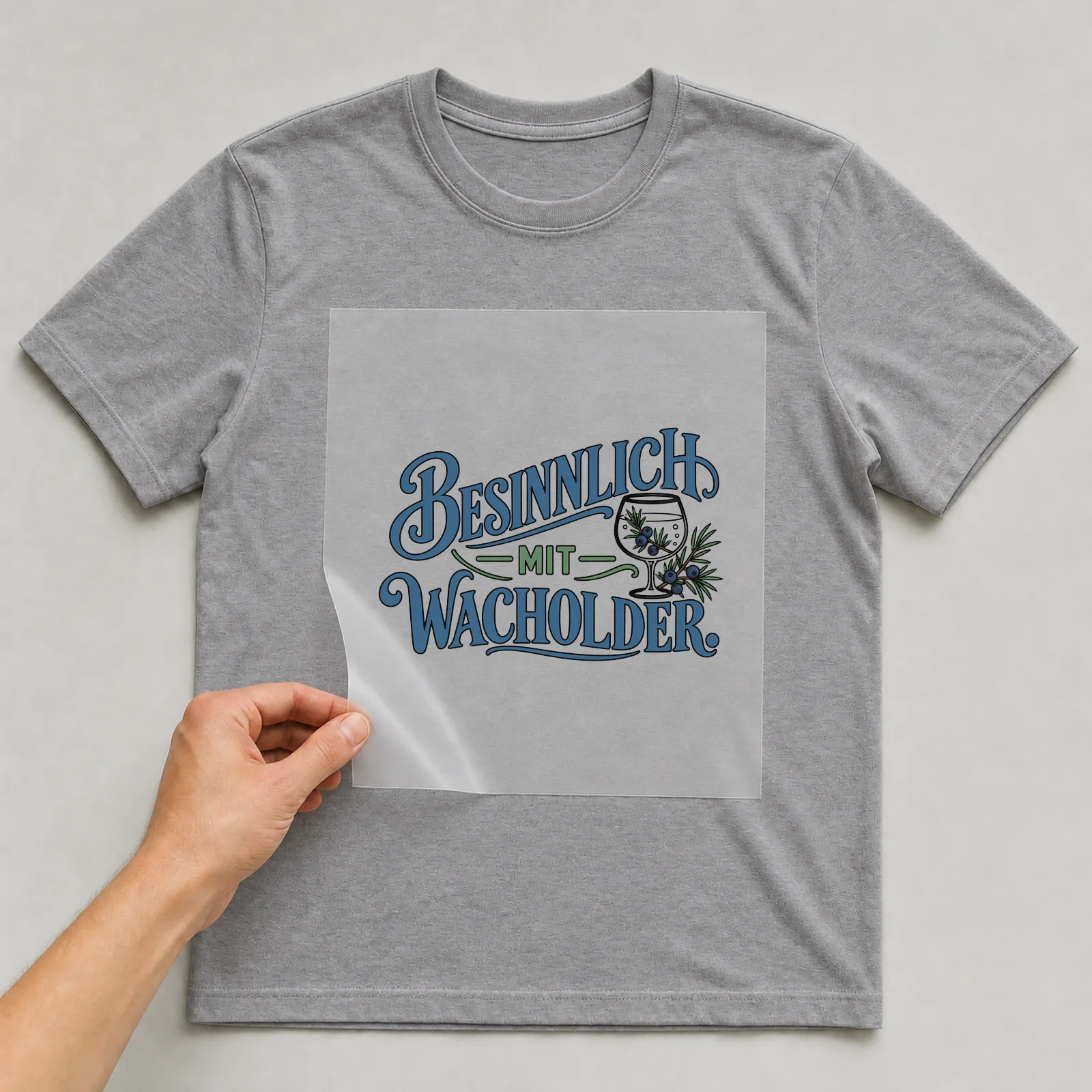 Bügelbild „Besinnlich mit Wacholder“ auf grauem T-Shirt, Folie wird aufgelegt
