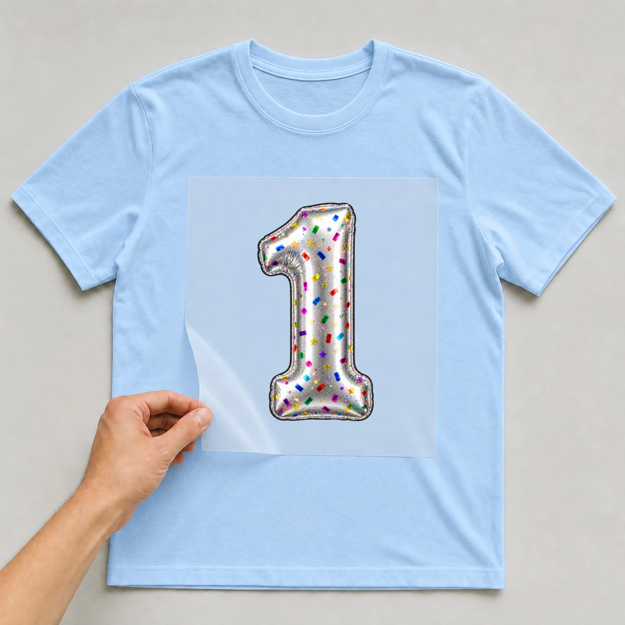 Bügelbild „Zahl 1 im Konfetti-Metallic-Look“ auf babyblauem T-Shirt, Folie wird aufgelegt