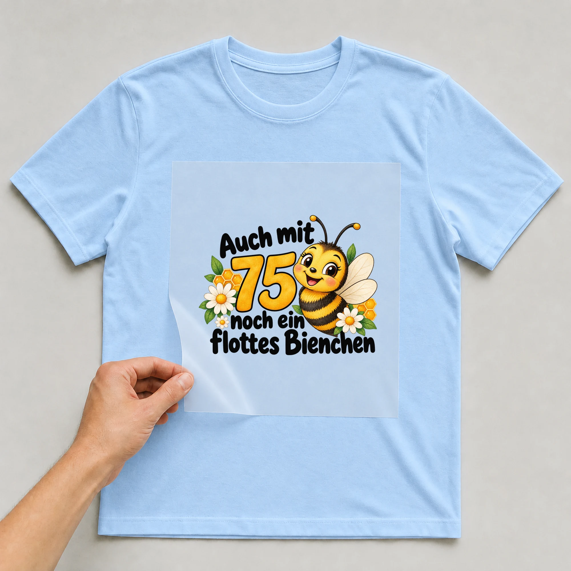 Bügelbild „Auch mit 75 noch ein flottes Bienchen“ auf babyblauem T-Shirt, Folie wird aufgelegt