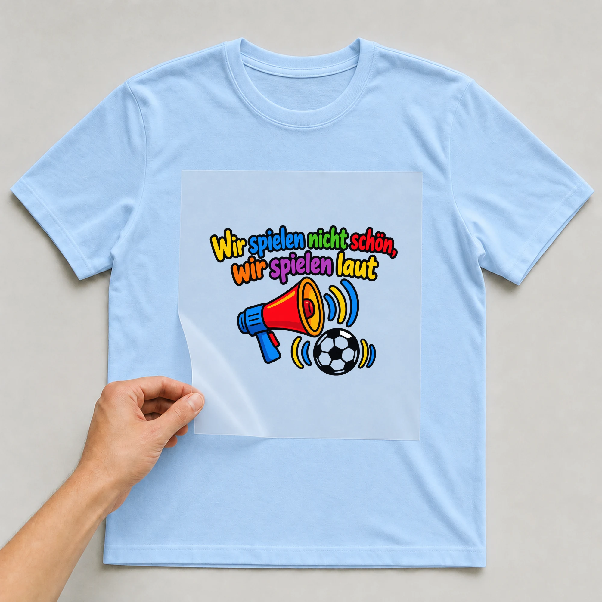 Bügelbild „Wir spielen nicht schön, wir spielen laut“ auf babyblauem T-Shirt, Folie wird aufgelegt