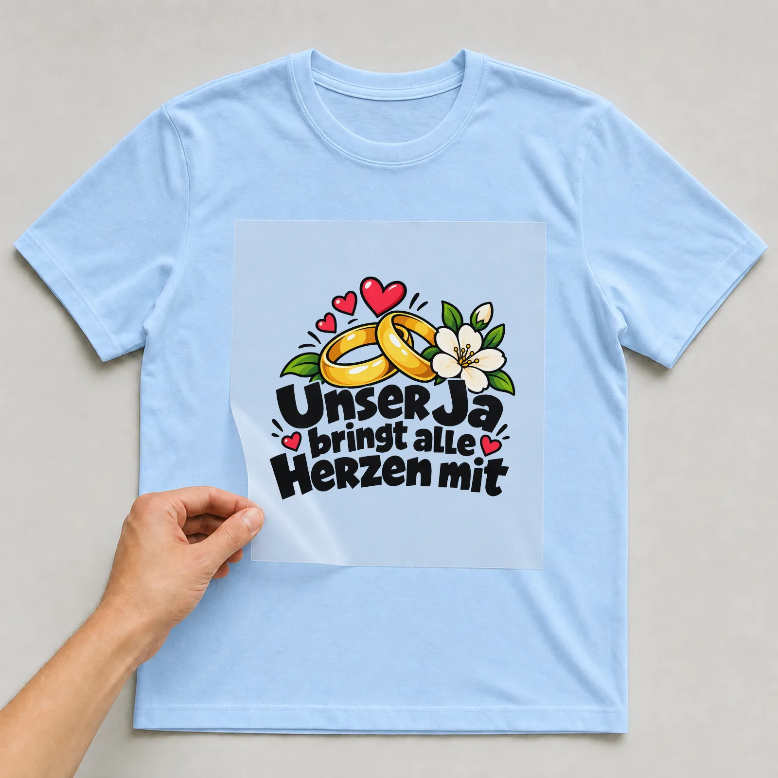 Bügelbild „Unser Ja bringt alle Herzen mit“ auf hellblauem T-Shirt, Folie wird aufgelegt