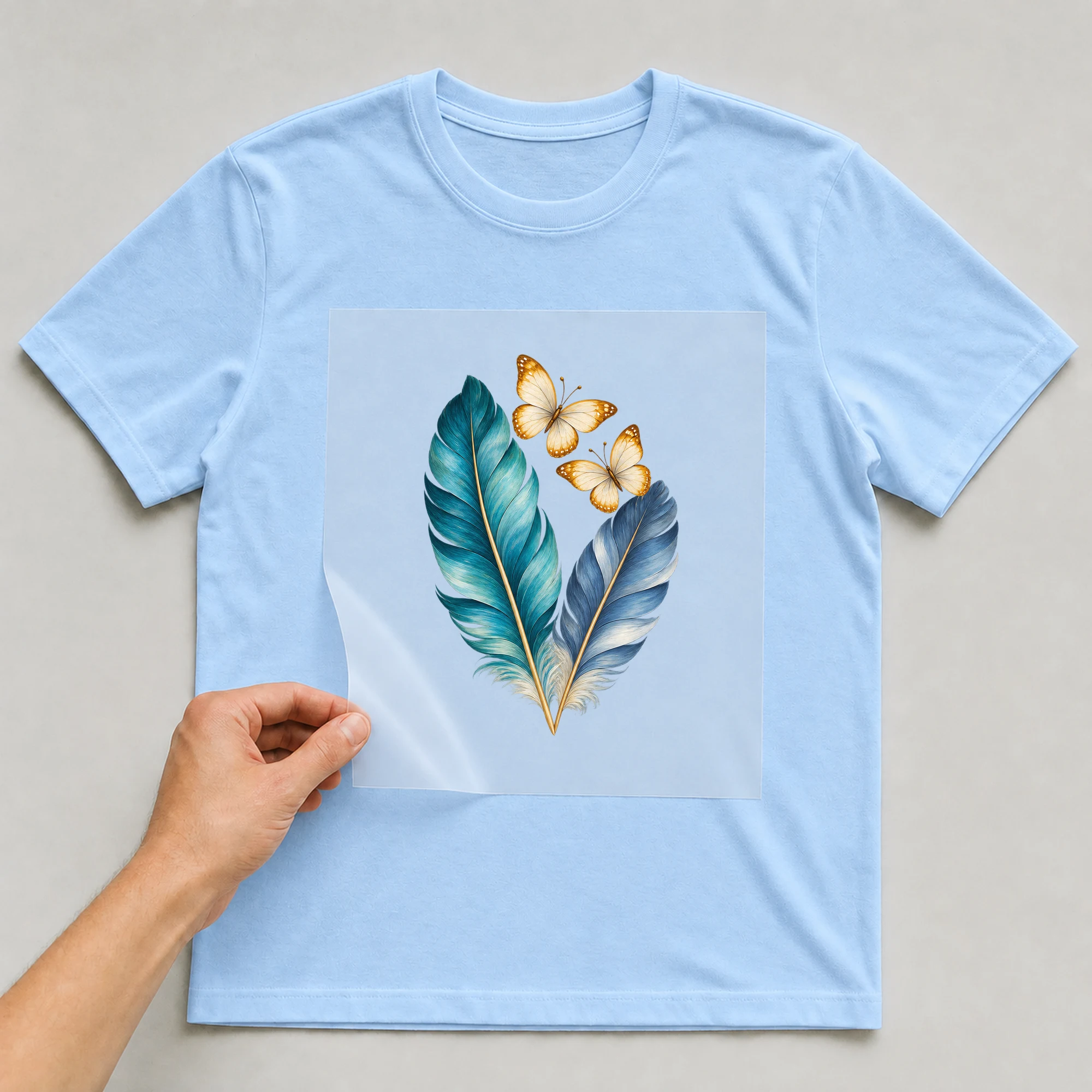 Bügelbild „Federn mit Schmetterlingen“ auf babyblauem T-Shirt, Folie wird aufgelegt