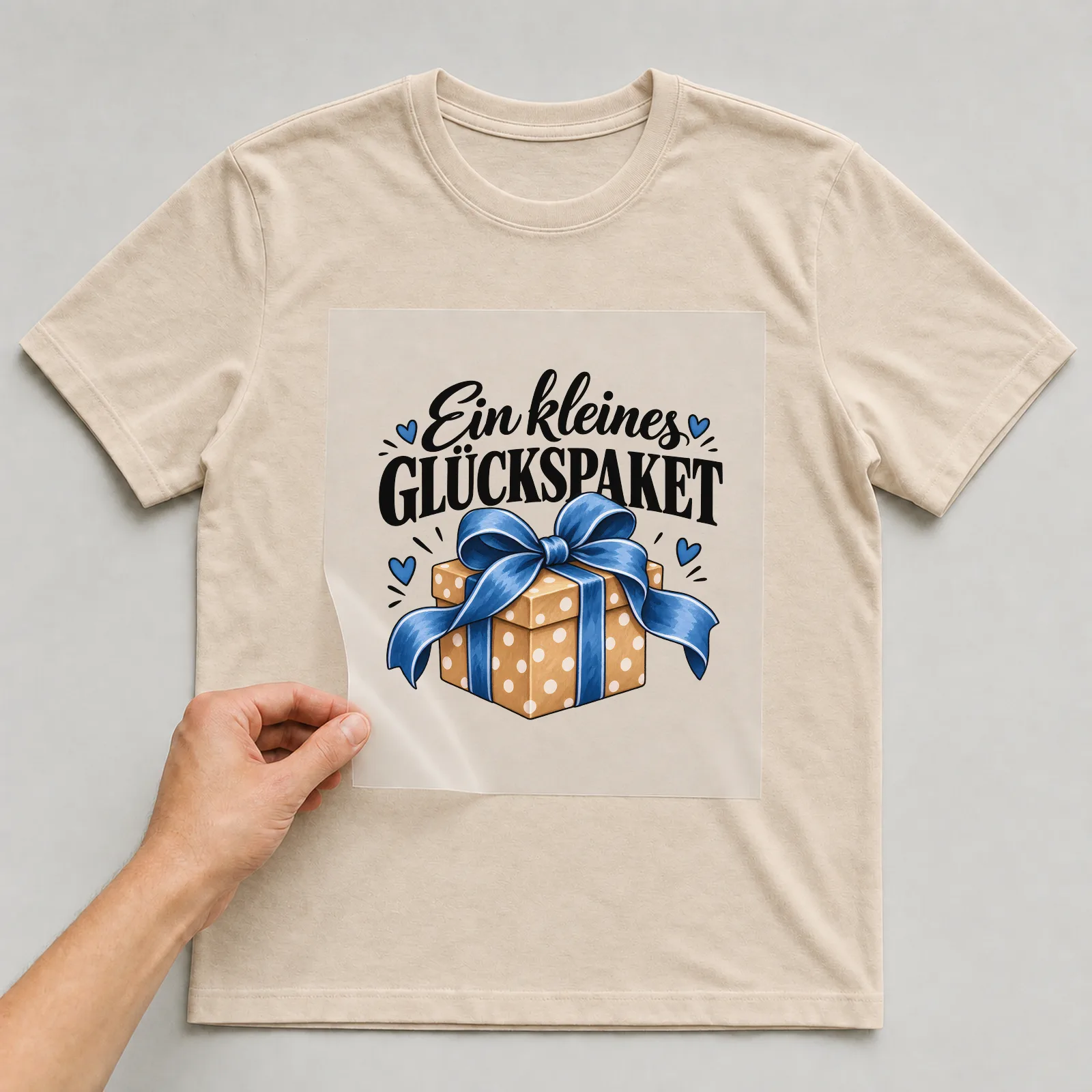 Bügelbild „Ein kleines Glückspaket“ auf beigem T-Shirt, Folie wird aufgelegt