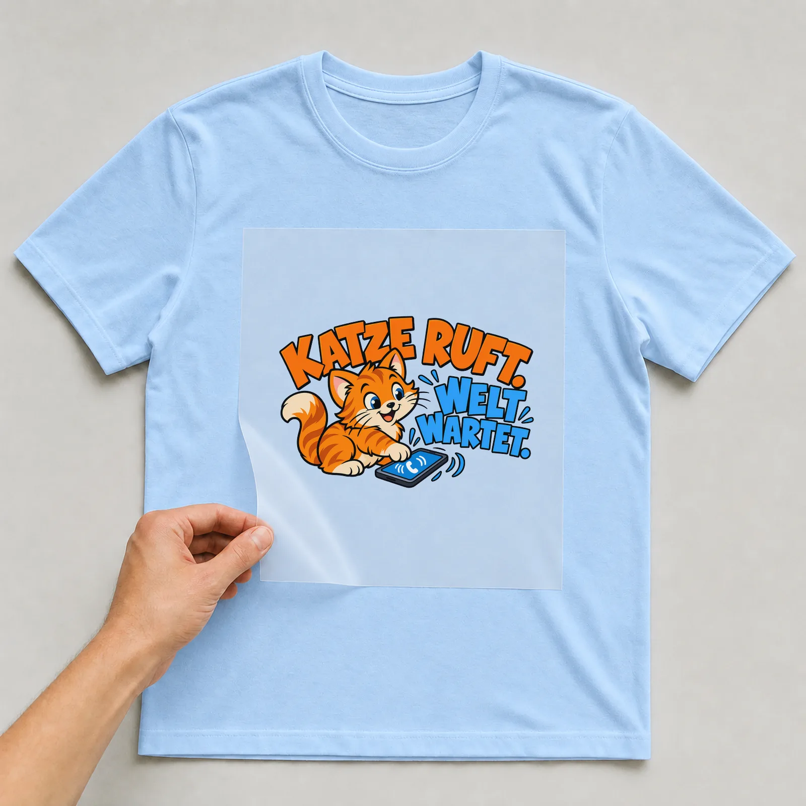 Bügelbild „Katze ruft. Welt wartet.“ auf hellblauem T-Shirt, Folie wird aufgelegt
