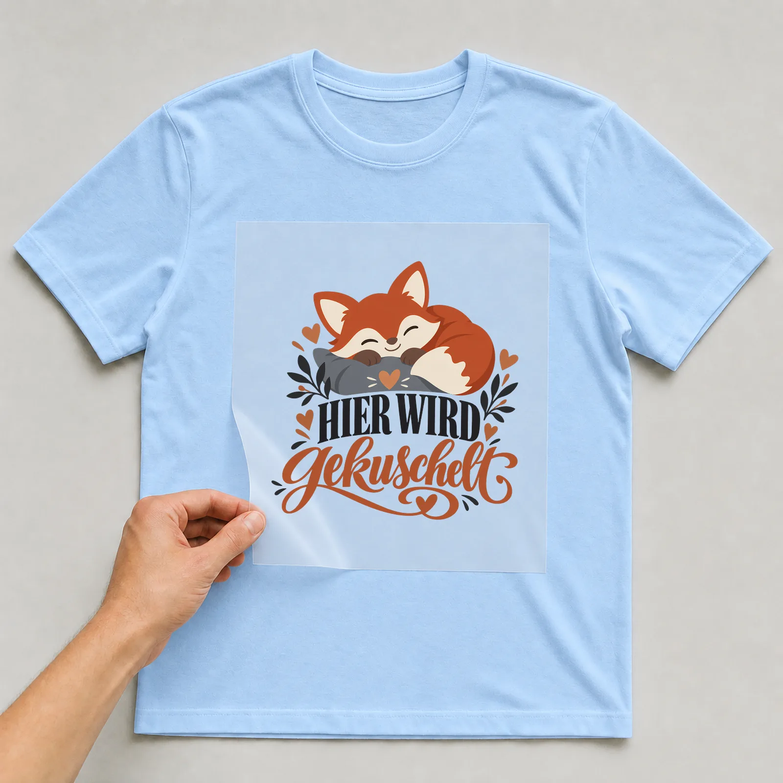 Bügelbild „Hier wird gekuschelt“ auf hellblauem T-Shirt, Folie wird aufgelegt