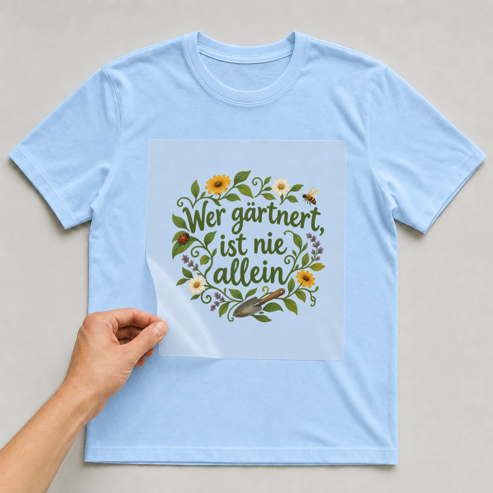 Bügelbild „Wer gärtnert, ist nie allein“ auf babyblauem T-Shirt, Folie wird aufgelegt
