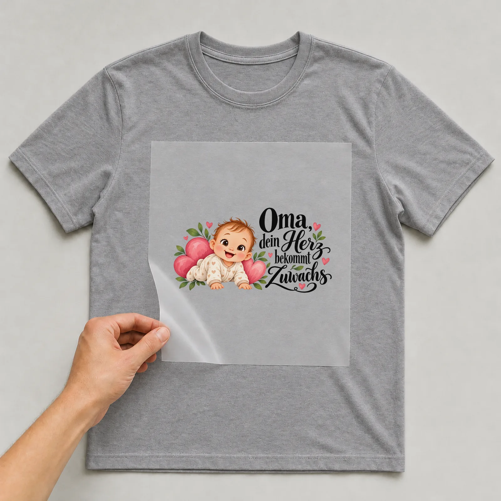 Bügelbild „Oma, dein Herz bekommt Zuwachs“ auf grauem T-Shirt, Folie wird aufgelegt