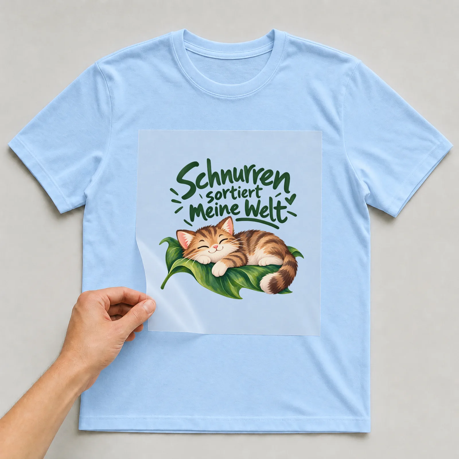 Bügelbild „Schnurren sortiert meine Welt“ auf hellblauem T-Shirt, Folie wird aufgelegt