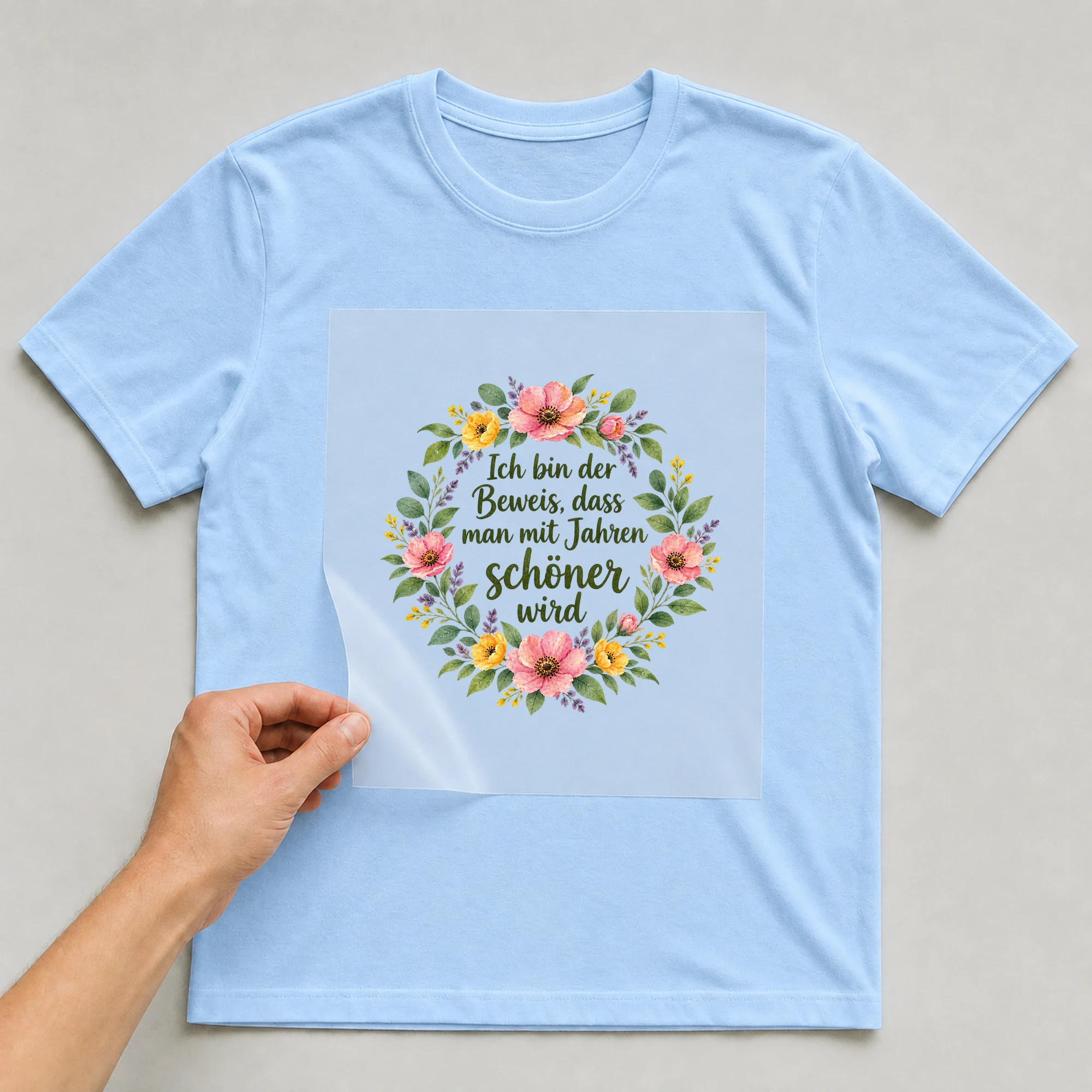 Bügelbild „Ich bin der Beweis, dass man mit Jahren schöner wird“ auf babyblauem T-Shirt, Folie wird aufgelegt