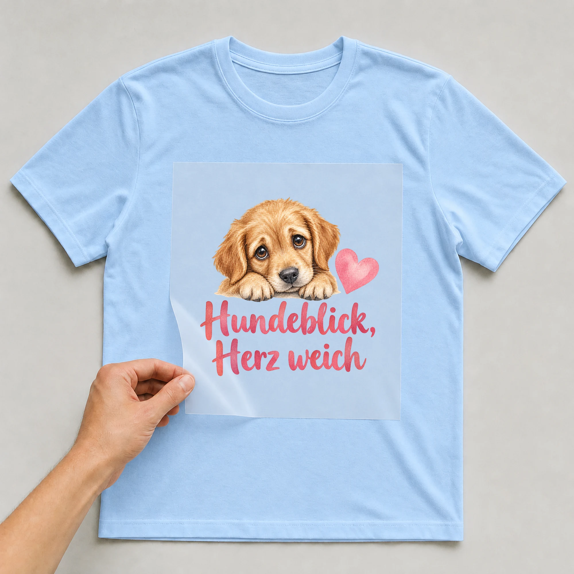 Bügelbild „Hundeblick, Herz weich“ auf babyblauem T-Shirt, Folie wird aufgelegt