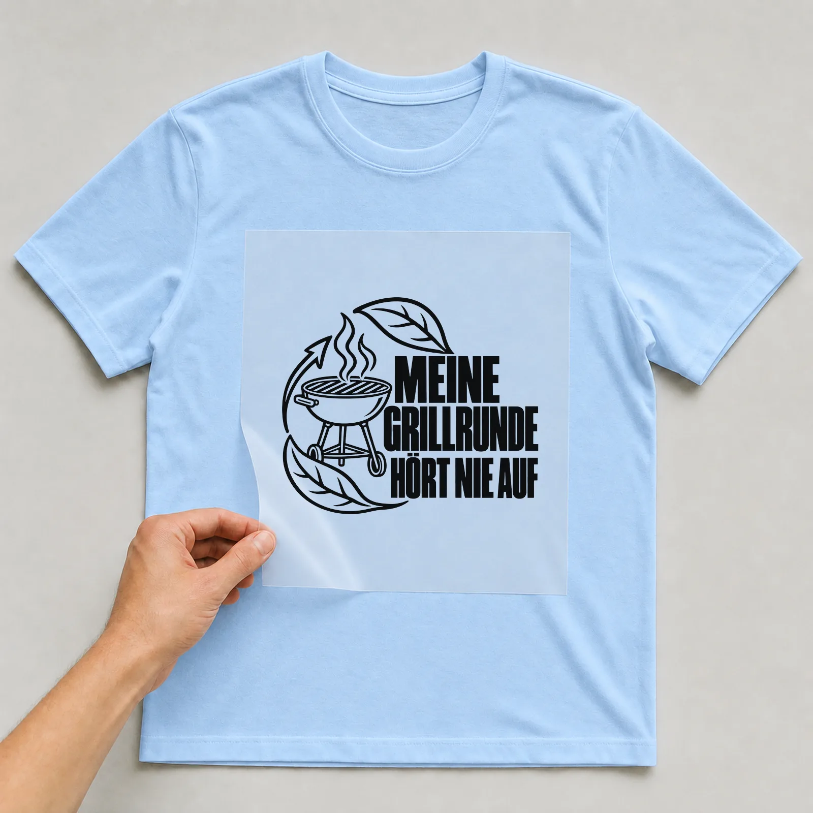 Bügelbild „Meine Grillrunde hört nie auf“ auf hellblauem T-Shirt, Folie wird aufgelegt