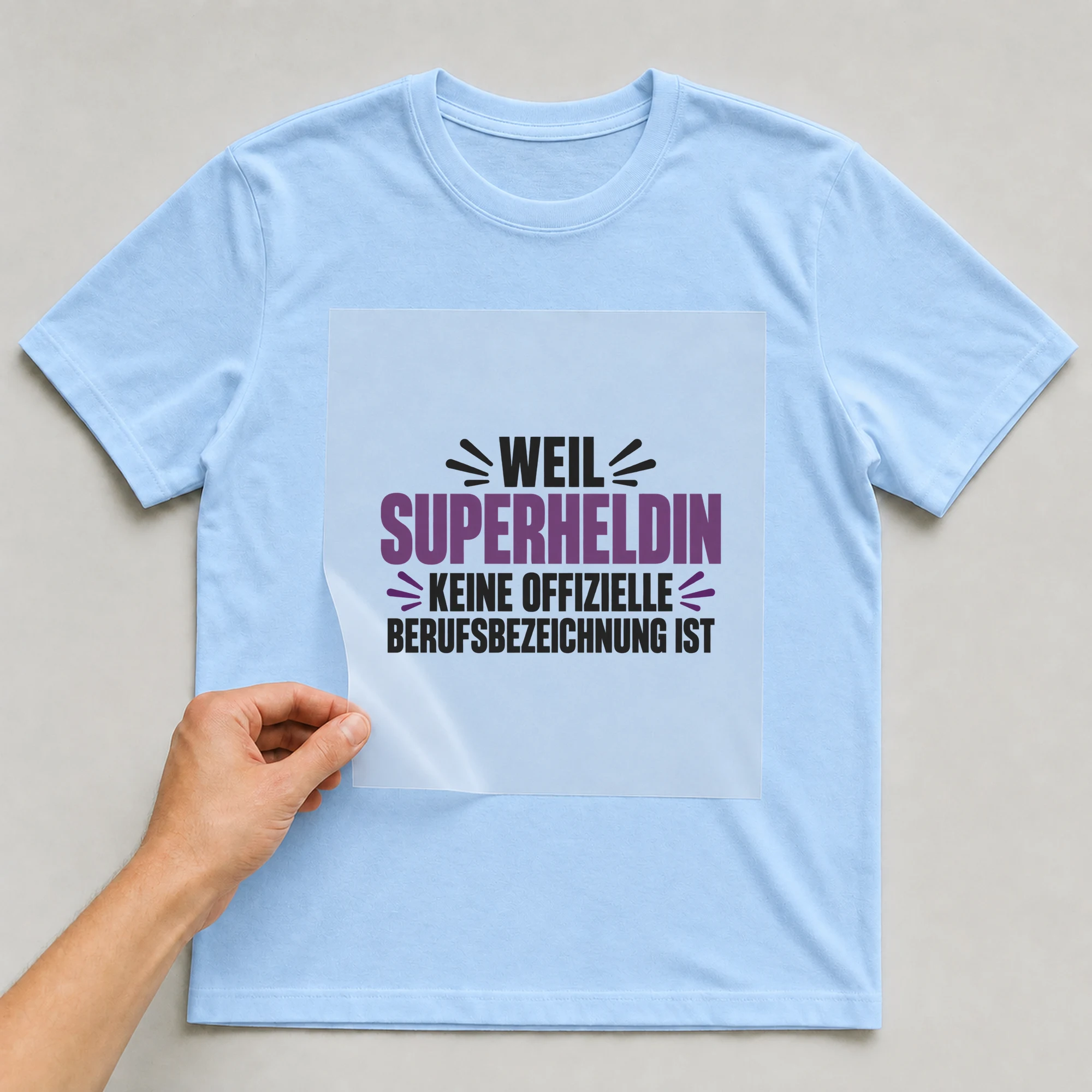 Bügelbild „Weil Superheldin keine offizielle Berufsbezeichnung ist“ auf babyblauem T-Shirt, Folie wird aufgelegt