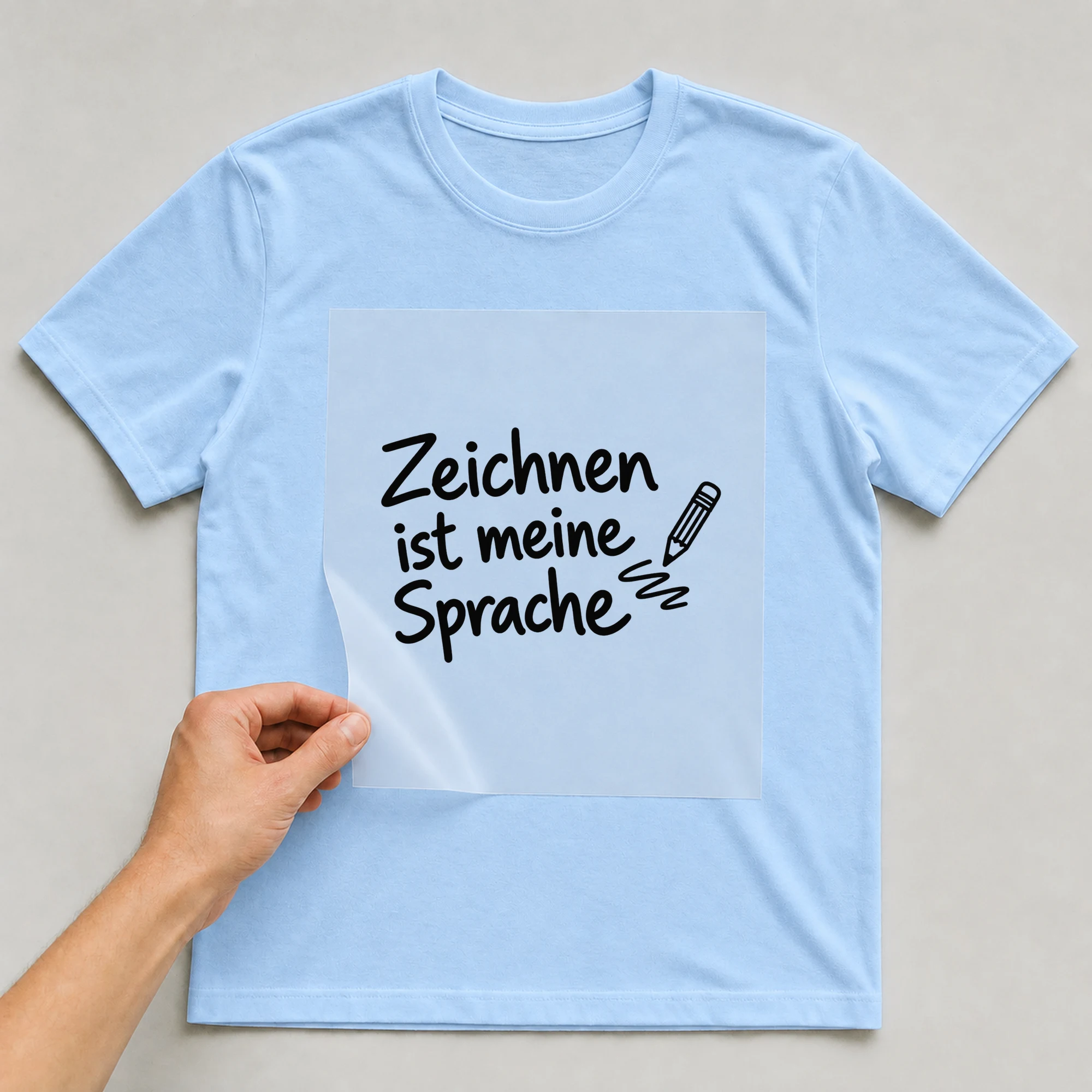 Bügelbild „Zeichnen ist meine Sprache“ auf babyblauem T-Shirt, Folie wird aufgelegt