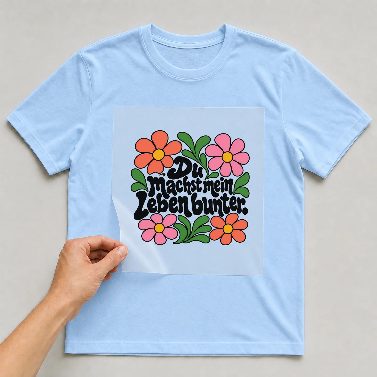 Bügelbild „Du machst mein Leben bunter“ auf hellblauem T-Shirt, Folie wird aufgelegt