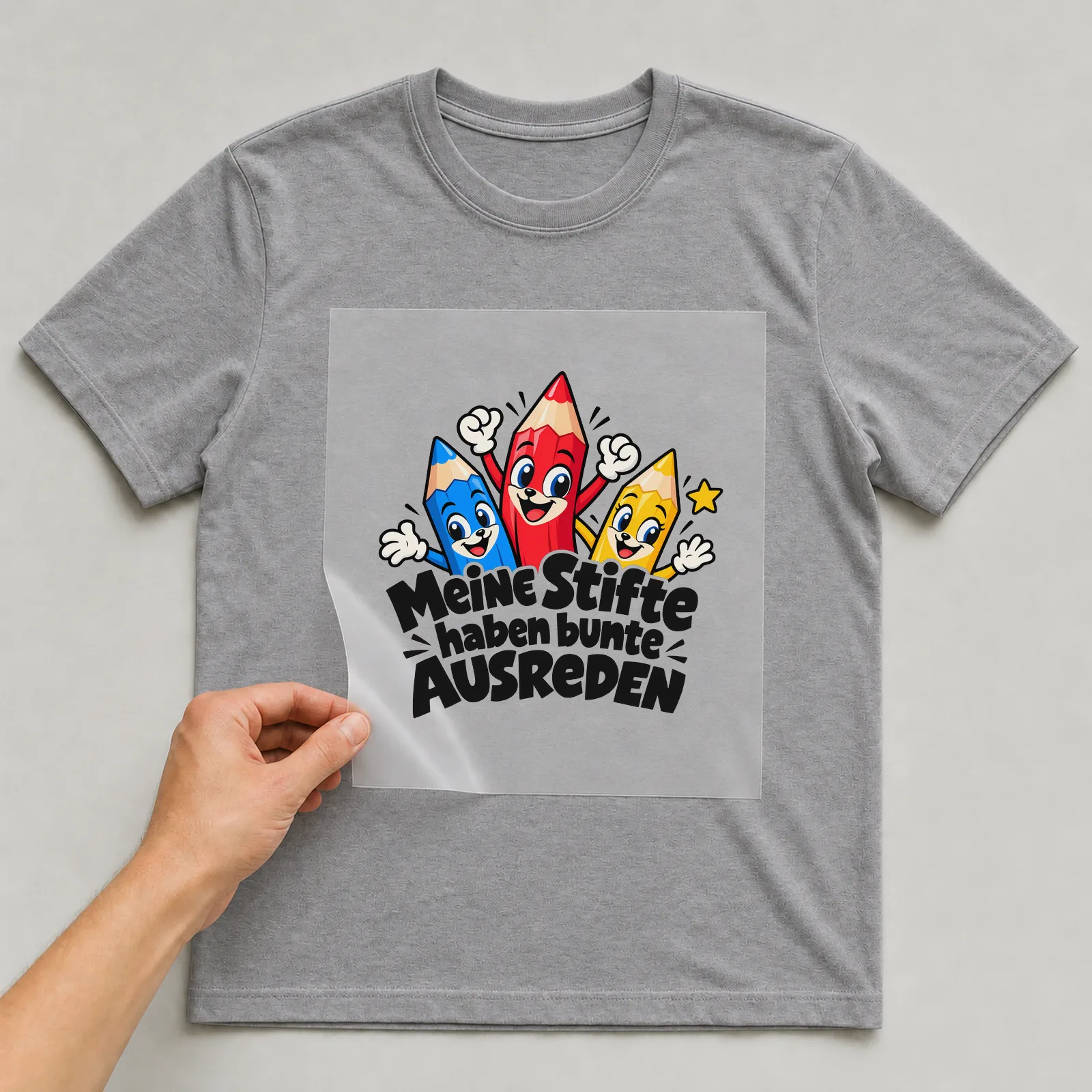 Bügelbild „Meine Stifte haben bunte AUSREDEN“ auf grauem T-Shirt, Folie wird aufgelegt
