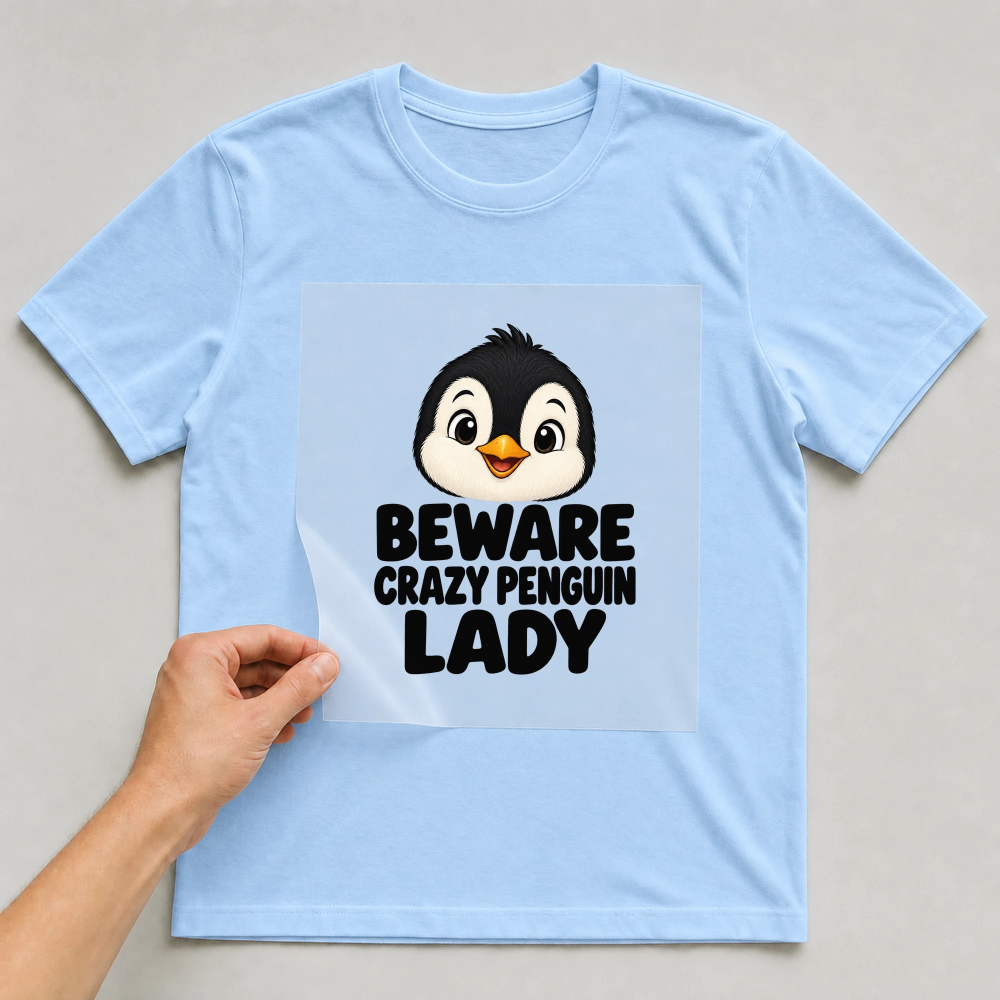 Bügelbild „Beware Crazy Penguin Lady“ auf babyblauem T-Shirt, Folie wird aufgelegt