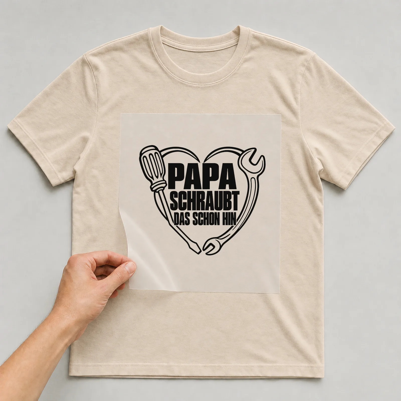 Bügelbild „Papa schraubt das schon hin“ auf beigem T-Shirt, Folie wird aufgelegt