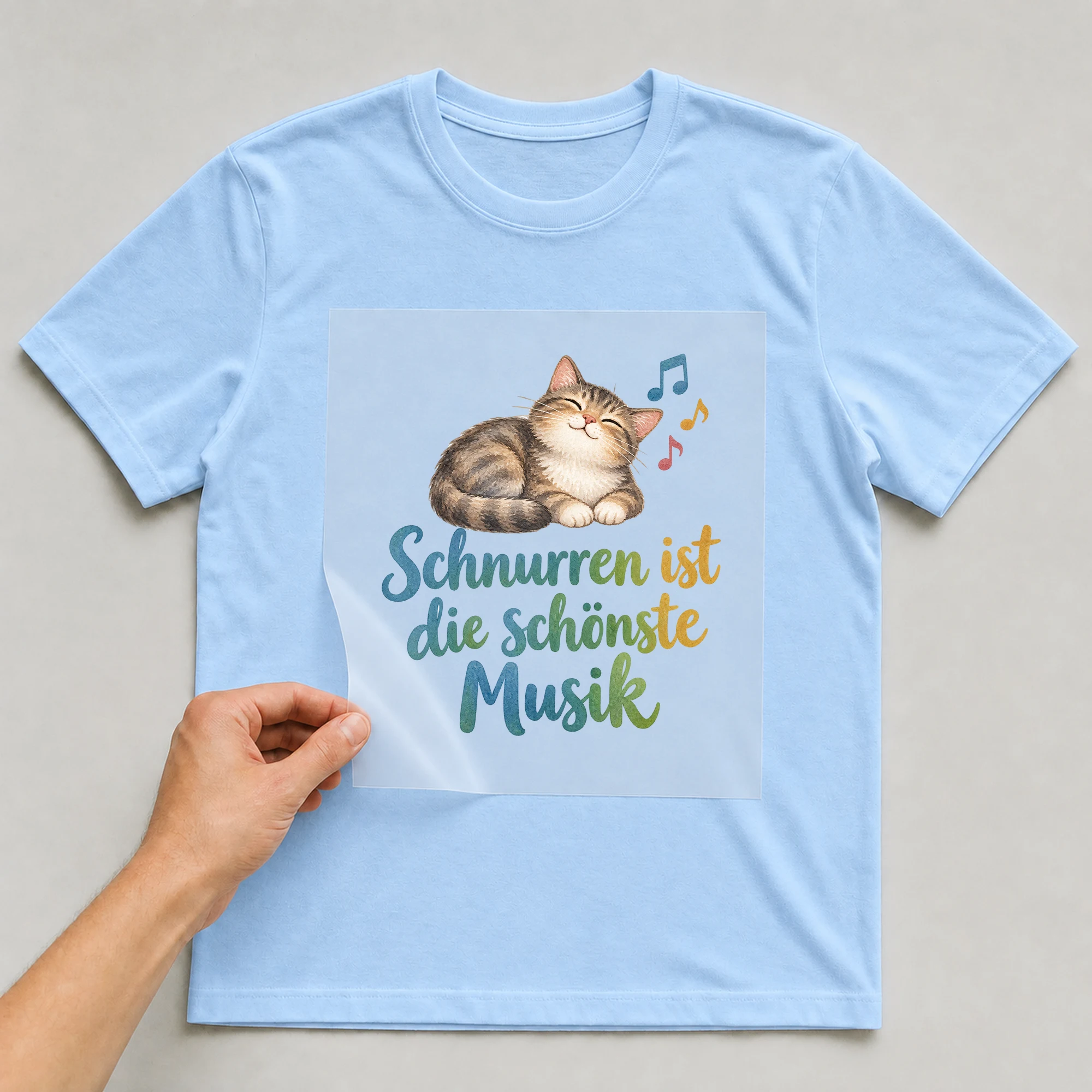 Bügelbild „Schnurren ist die schönste Musik“ auf babyblauem T-Shirt, Folie wird aufgelegt