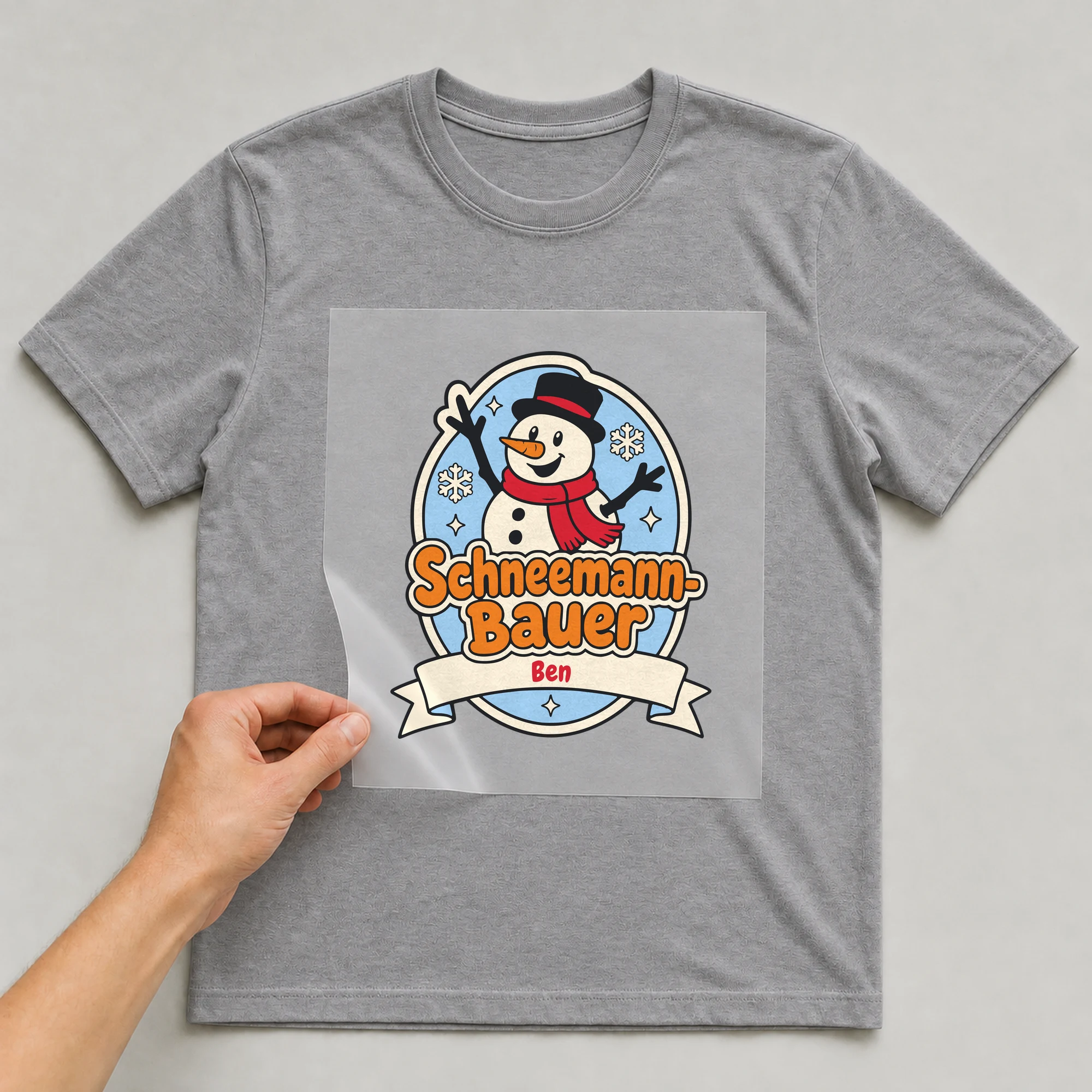 Bügelbild Schneemann-Bauer mit Name auf grauem T-Shirt, Folie wird aufgelegt