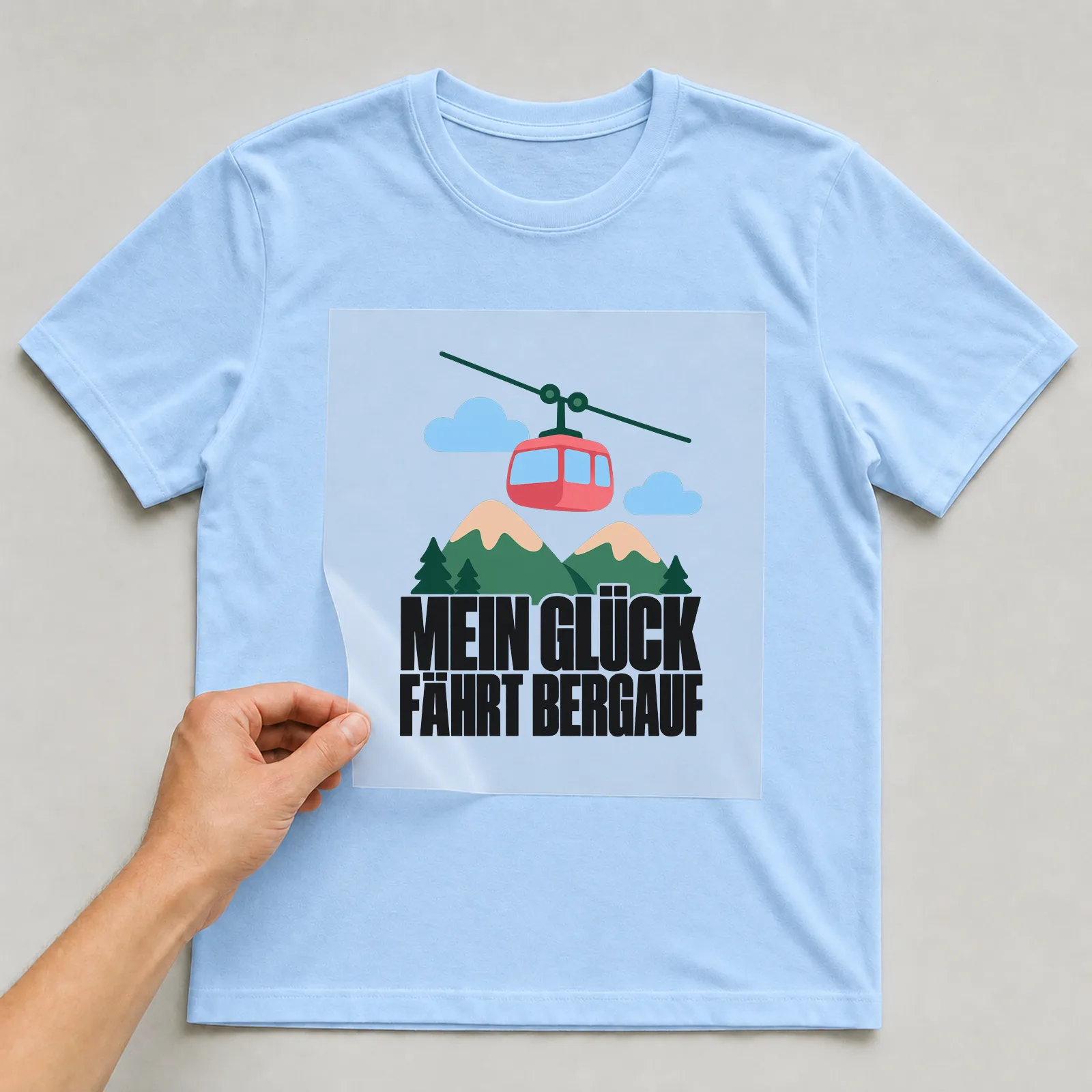 Bügelbild „Mein Glück fährt bergauf“ auf hellblauem T-Shirt, Folie wird aufgelegt