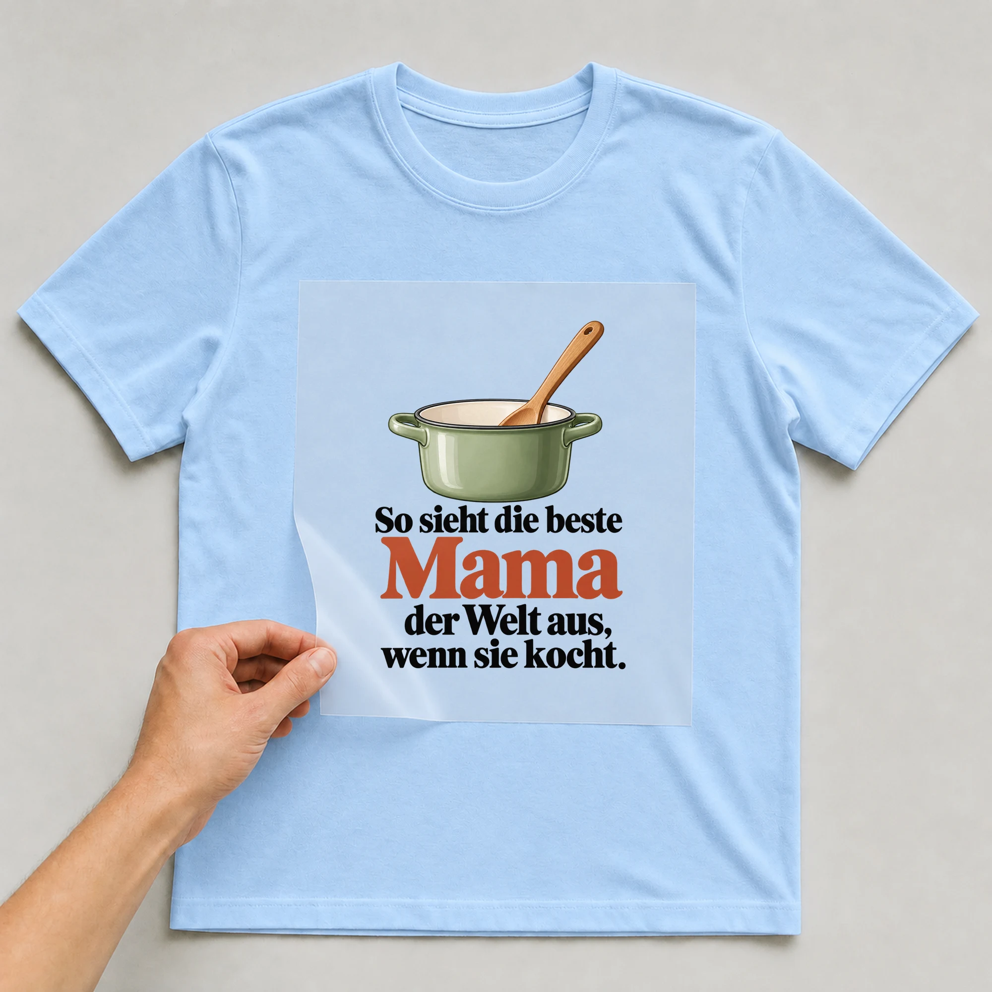 Bügelbild „So sieht die beste Mama der Welt aus, wenn sie kocht.“ auf babyblauem T-Shirt, Folie wird aufgelegt