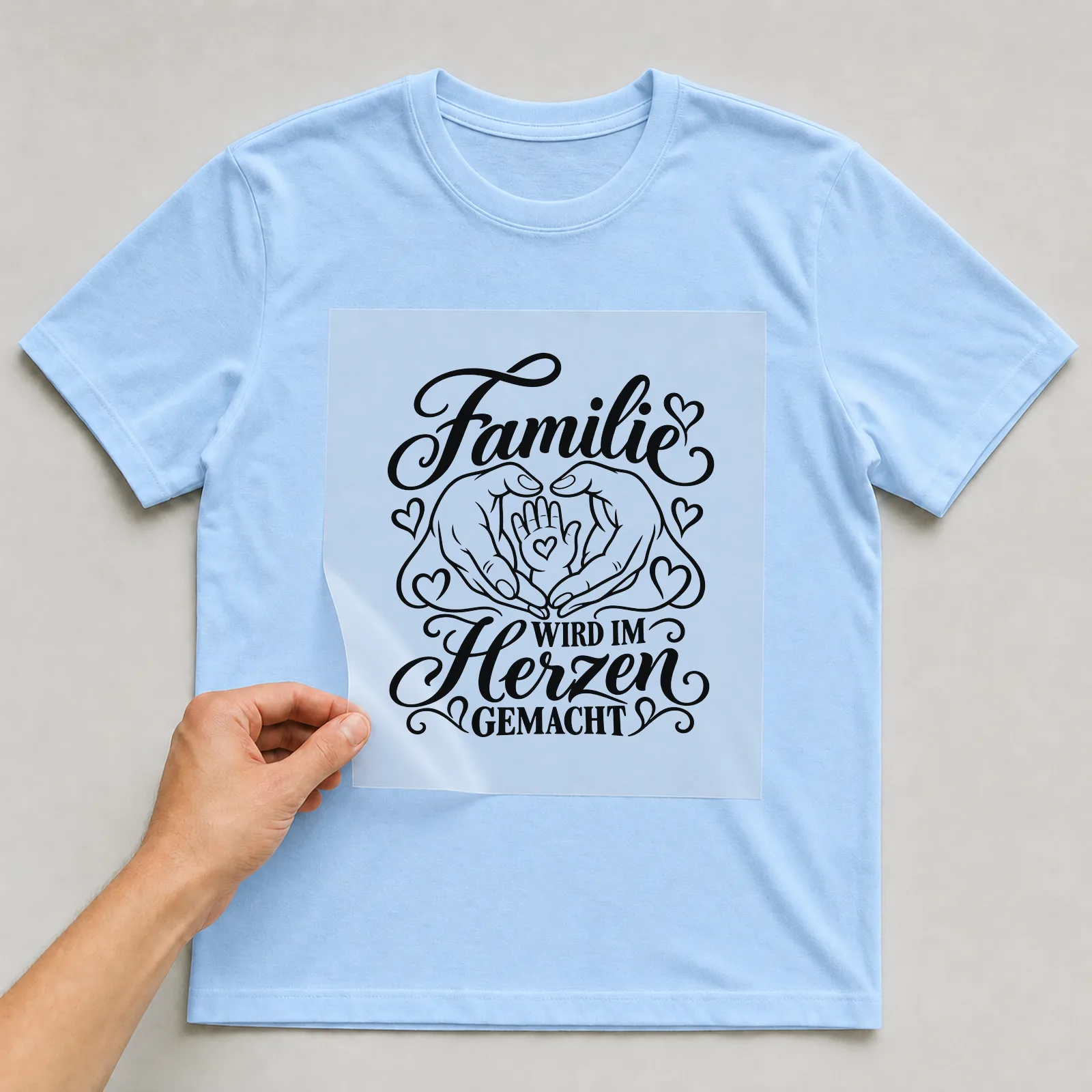 Bügelbild „Familie wird im Herzen gemacht“ auf hellblauem T-Shirt, Folie wird aufgelegt